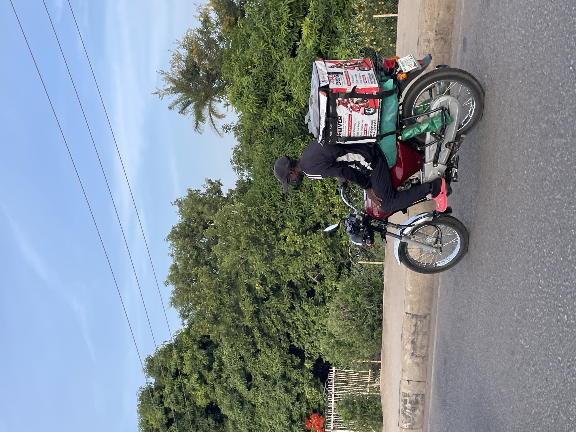 The image shows a delivery person on a red motorcycle in Jos, Nigeria. The rider, wearing a black cap, black jacket with a white stripe, and black pants, is straddling the motorcycle. A delivery box branded with "FLIVER" is secured on the back, containing visible phone numbers 8030955580 and 704-077339. The motorcycle is parked on the side of a paved road. Lush green foliage and trees are in the background, beneath a clear, blue sky. Overhead power lines are visible in the sky.