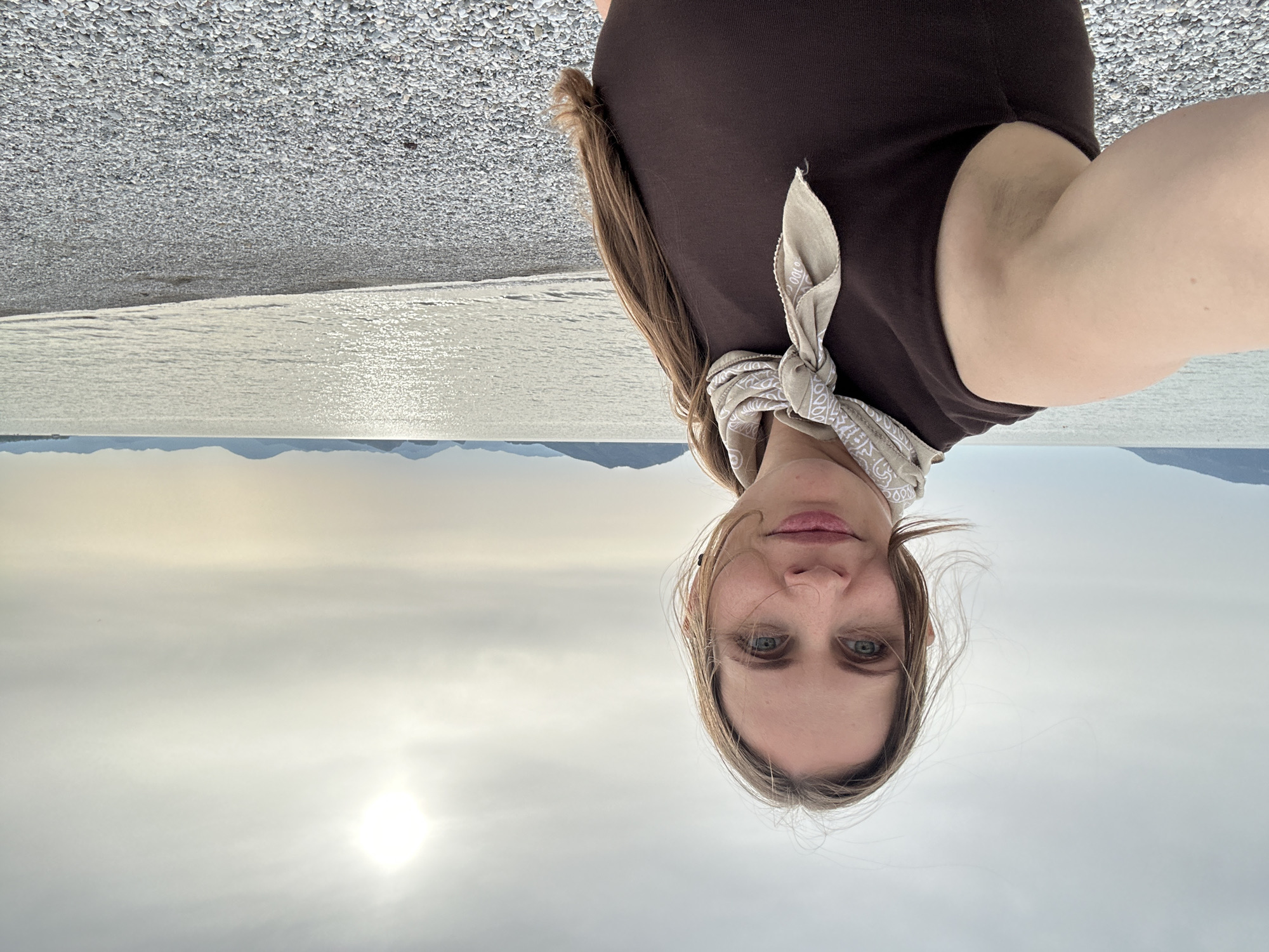 The image is a selfie of a woman, rotated upside down, taken outdoors near a body of water, likely on a beach. The woman has light skin, light brown hair, and blue eyes. She is wearing a brown top and a light-colored bandana around her neck. The background shows a beach covered in small white stones or shells, a body of water with a reflection of the sun, and distant mountains. The sky is overcast. The location is potentially in Fethiye, Turkey, given the landscape.