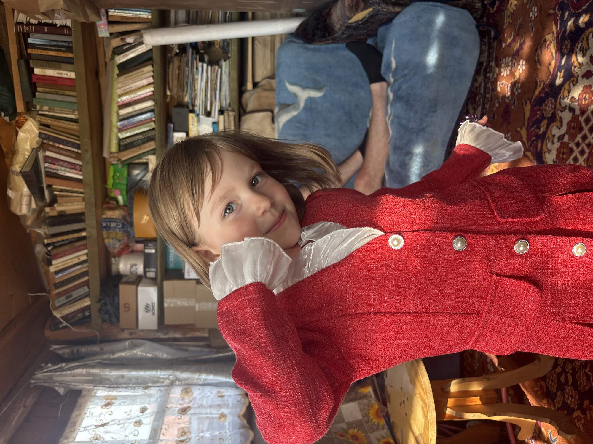A young girl with blonde hair and blue eyes stands in a cluttered room, wearing a red jacket with white ruffles. She is positioned in the foreground, facing the camera with her head tilted and her right hand near her face. Behind her, bookshelves filled with books and various items line the walls. A blue blanket with a white bird-like pattern covers a sofa on which a person's legs are visible. The room appears to be a storage area or a casual living space. The lighting suggests it is daytime, with sunlight streaming in from a window to the left. The overall impression is of an indoor, informal setting. The specific location in the city of Маркидоновка, Russia is not determinable from the visual information in the image.