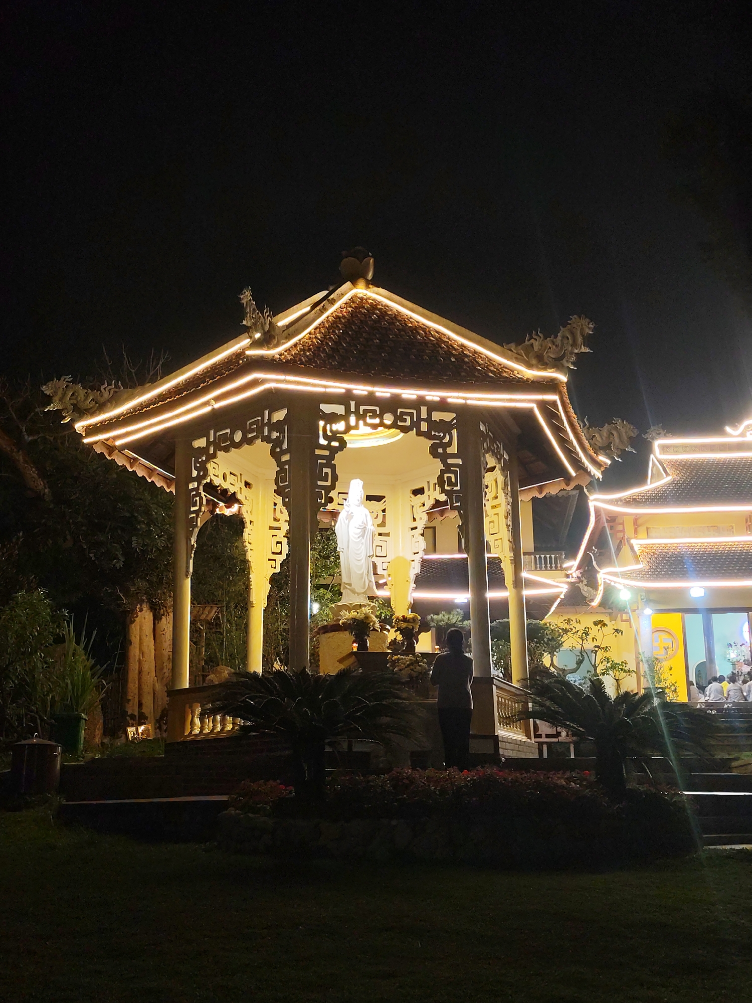 The image depicts a serene nighttime scene at a temple or pagoda, likely in Da Lat, Vietnam, as suggested by the context.
In the foreground, a brightly lit, ornate pavilion stands centrally. Its multi-tiered, tiled roof is extensively outlined with warm string lights, and its edges feature intricate, dragon-like sculptures. Inside the pavilion, a prominent white statue, resembling a Bodhisattva (possibly Guan Yin or Kuan Yin), is illuminated, with flower offerings placed at its base. A single individual, seen from behind, stands before the statue, seemingly in contemplation or prayer.
To the right, another larger temple building shares a similar architectural style, with its rooflines also adorned with warm string lights and decorative sculptures. This building features a lighter-colored facade, possibly yellow, and a circular symbol (resembling a Buddhist swastika, a symbol of auspiciousness) is faintly visible on its wall. A group of people are gathered on steps or platforms in front of this building, suggesting a communal activity or religious service.
The surrounding environment is dark but appears lush, with various trees, bushes, and potted plants framing the structures. A grassy area occupies the immediate foreground. The night sky is deep black, allowing the artificial lights to glow brightly and define the architectural details. The overall ambiance is one of peace and reverence. No clear, legible text is visible in the image.