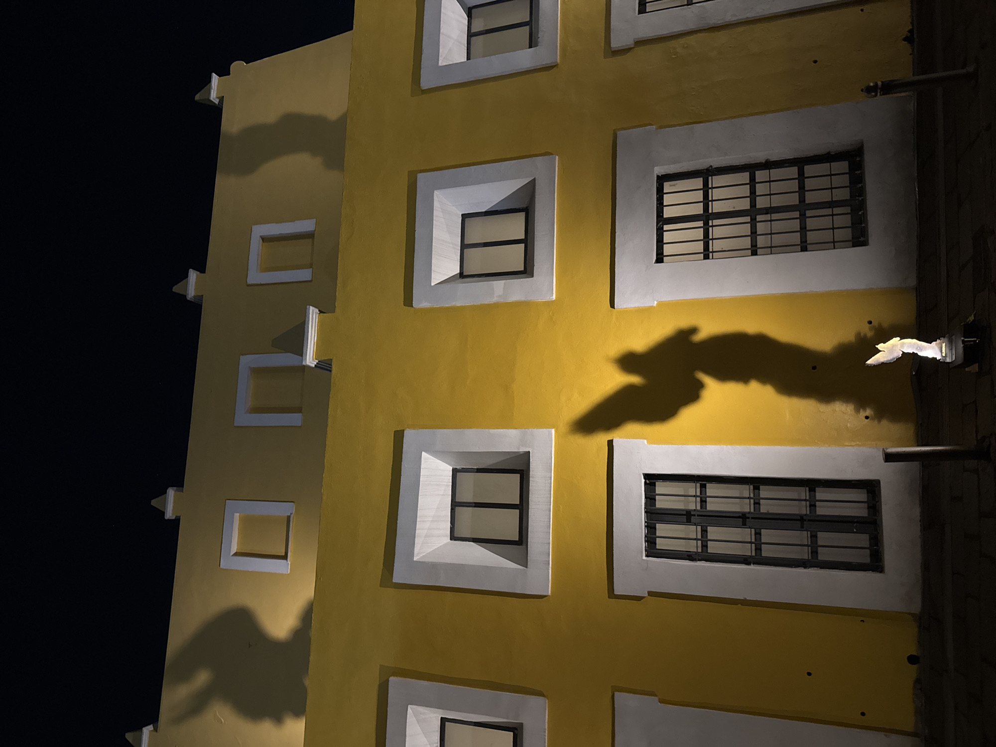 A nighttime exterior shot of a yellow, multi-story building in Puebla, Mexico. The building has rectangular windows with white frames. Some windows have metal bars. Shadows cast on the building suggest foliage or structures nearby. The top left shows only a sliver of sky which is black. The building is angled to the left. A pipe is visible on the right side of the building.