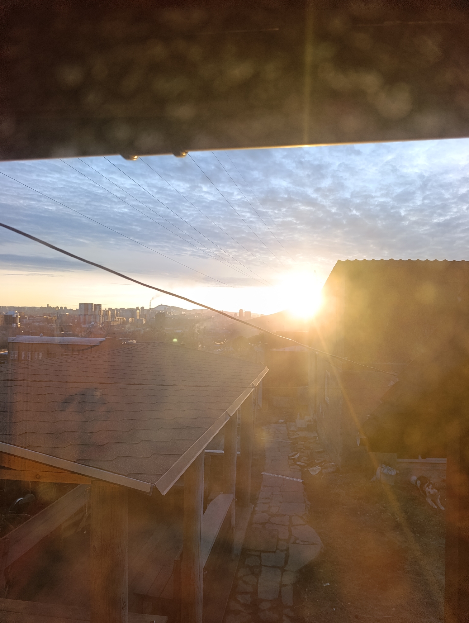 The image shows a view from a window or opening overlooking a city landscape at sunrise or sunset. The presence of a mountain with a smokestack in the distance suggests the location may be Krasnoyarsk, Russia, as the city is known for its industrial presence and surrounding natural features. A low-rise building with a tiled roof and a wooden structure beneath it, perhaps a veranda or pergola, is visible in the foreground. There is a narrow, paved or stone-lined pathway running alongside the building. On the right side of the image, adjacent to the pathway, a dog is lying down in the sun. Overhead power lines run across the sky. The sky is cloudy with the sun obscured by the haze, but the bright light indicates it is near the horizon.