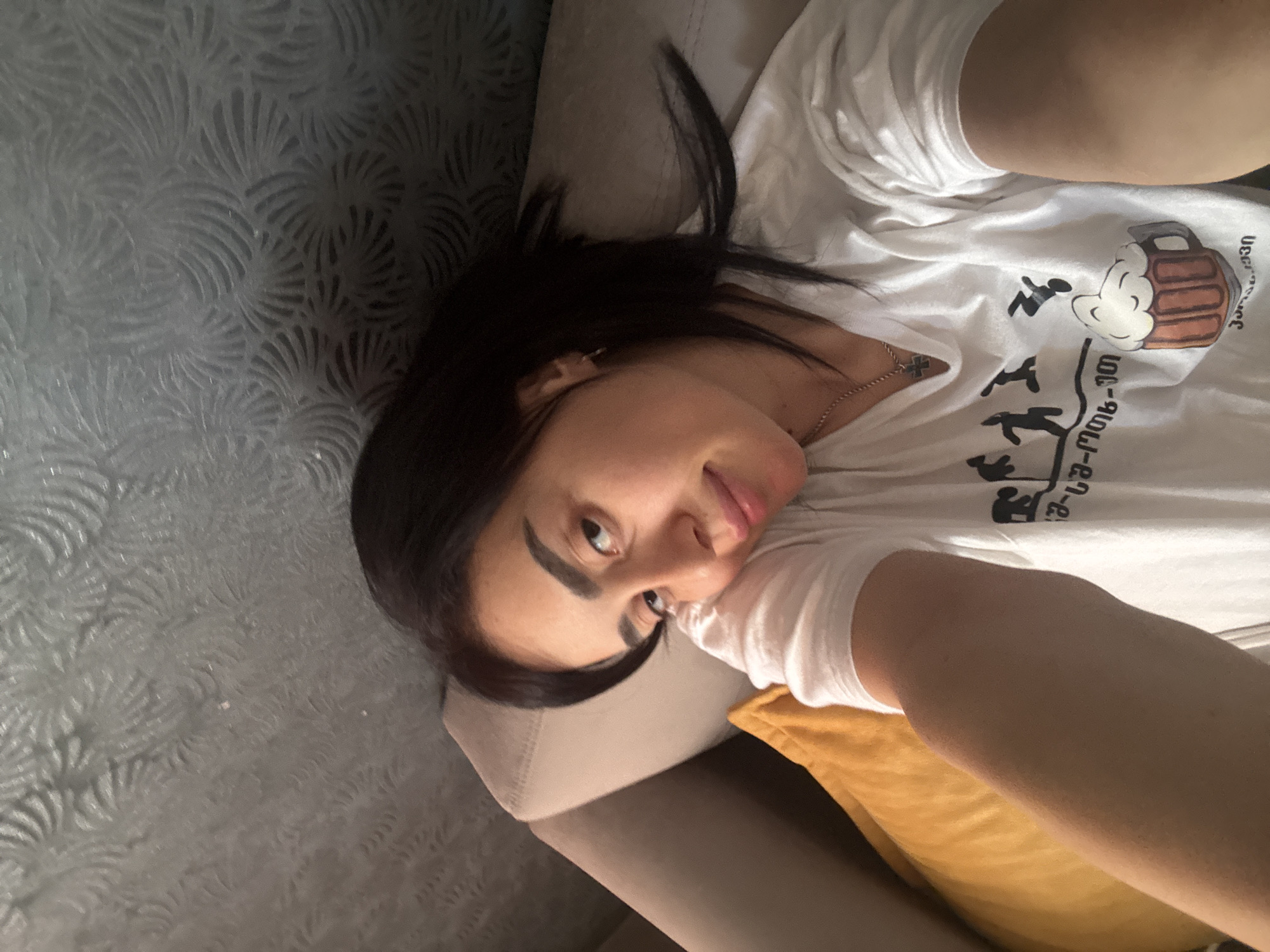 A woman lies on a couch against a patterned wall. She is wearing a white T-shirt with a graphic featuring cartoon figures and a beer mug, with text in what appears to be Georgian script. The scene is indoors, likely in a residence in Tbilisi, Georgia, based on the Georgian text. The woman is looking at the camera. A mustard yellow pillow is visible beside her on the couch. The wall behind the couch is decorated with a repeating floral or leaf-like pattern.
