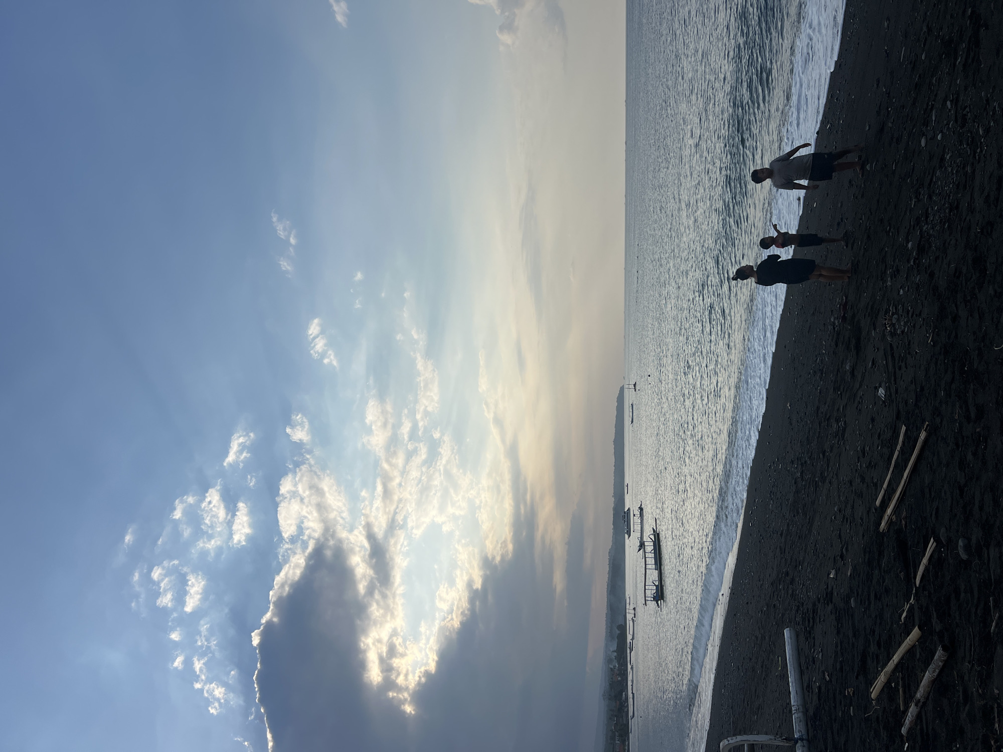 In the city of Abang, Indonesia, a serene beach scene unfolds. The sky above is a canvas of blues, dotted with scattered clouds, some illuminated by the setting sun. Rays of light pierce through the clouds, casting a soft glow over the horizon. The ocean stretches out, its surface rippling with gentle waves that kiss the dark, volcanic sand of the beach. A few traditional boats, some with thatched roofs, are anchored offshore, hinting at the local fishing or tourism activities.
On the beach, three figures are silhouetted against the sea and sky. Two children and an adult stand near the water's edge, their forms dark and indistinct in the dimming light. One child points towards the sea, perhaps at a boat or a marine creature. The other figures appear to be observing the water or the child's gesture. Scattered across the dark sand are pieces of driftwood and bamboo, remnants of the sea's activity or perhaps materials for crafting. The overall atmosphere is one of tranquility and natural beauty, a moment captured at the cusp of day and night.