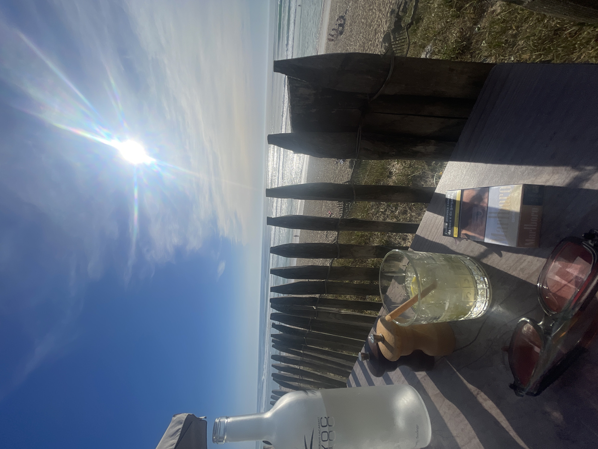 The image shows a beach scene in Lacanau, France, with a focus on a table setting. On the table are a bottle of "88" liquor, a glass of a clear beverage with a straw and lemon, a pack of Marlboro cigarettes, and a pair of sunglasses. In the background, a wooden fence separates the table from the beach. The beach itself has people visible in the distance, and the sky is bright with the sun.