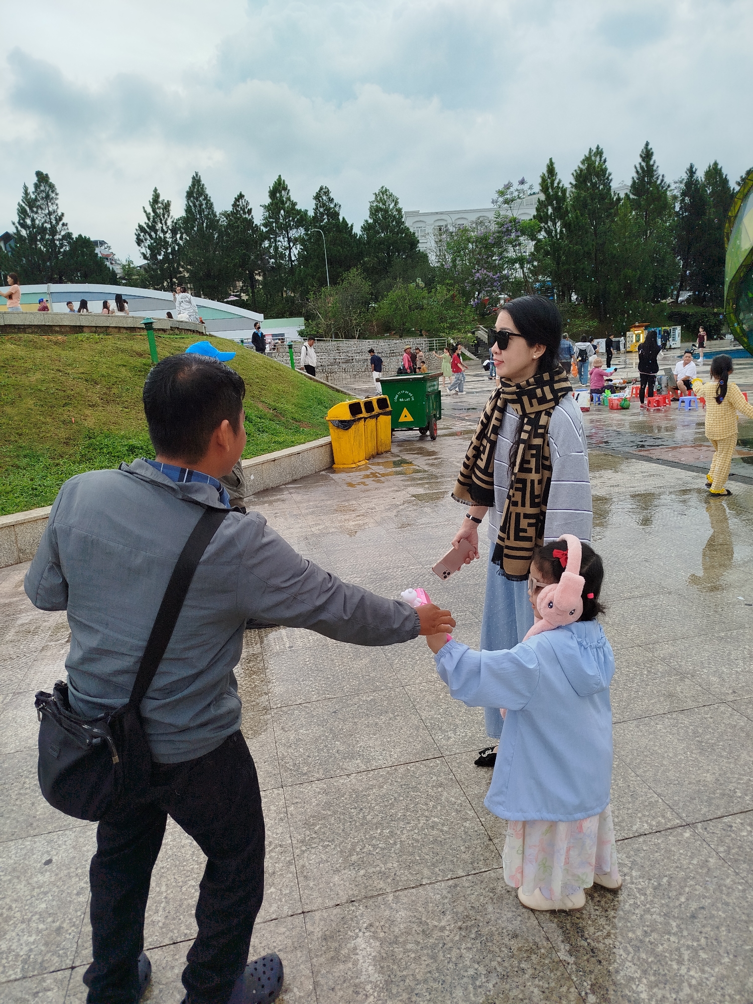 The scene is an outdoor public area in Da Lat, Vietnam, likely a park or plaza based on the presence of trees and the presence of people. The ground is paved with large, light-colored stones, and puddles indicate it has recently rained. A man in a gray jacket and black pants faces away from the camera, holding a pink toy and the hand of a child wearing a light blue jacket, floral skirt, and pink earmuffs. A woman, possibly the child's mother, stands next to the child wearing sunglasses and a patterned scarf. In the background, there are other people, trees, buildings, and some public amenities like trash cans. The weather appears overcast.