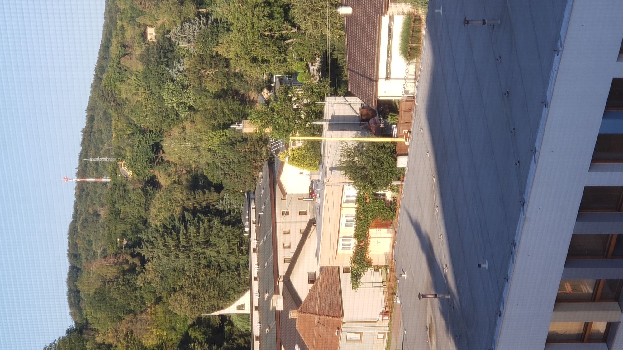 The image is a high-angle view looking down and across a town, featuring a mix of residential and industrial buildings, surrounded by a heavily wooded hillside. In the background, a red and white telecommunications tower stands on the crest of the hill, accompanied by a metal electrical pylon. The foreground shows the flat, grey rooftop of a modern building with visible ventilation pipes and mounting fixtures, and shadows cast across it. Below this rooftop are several other buildings with varying roof styles, including orange tile and grey flat roofs, and light-colored facades. Some buildings have prominent windows and chimneys. Dense green trees dominate the middle and background, with some showing signs of autumn color. A white steeple of a church or religious building is visible towards the upper left. The sky is clear and blue. The scene suggests a suburban or semi-rural area on a sunny day. The location is the area of Fintice, in the vicinity of Prešov, Slovakia.