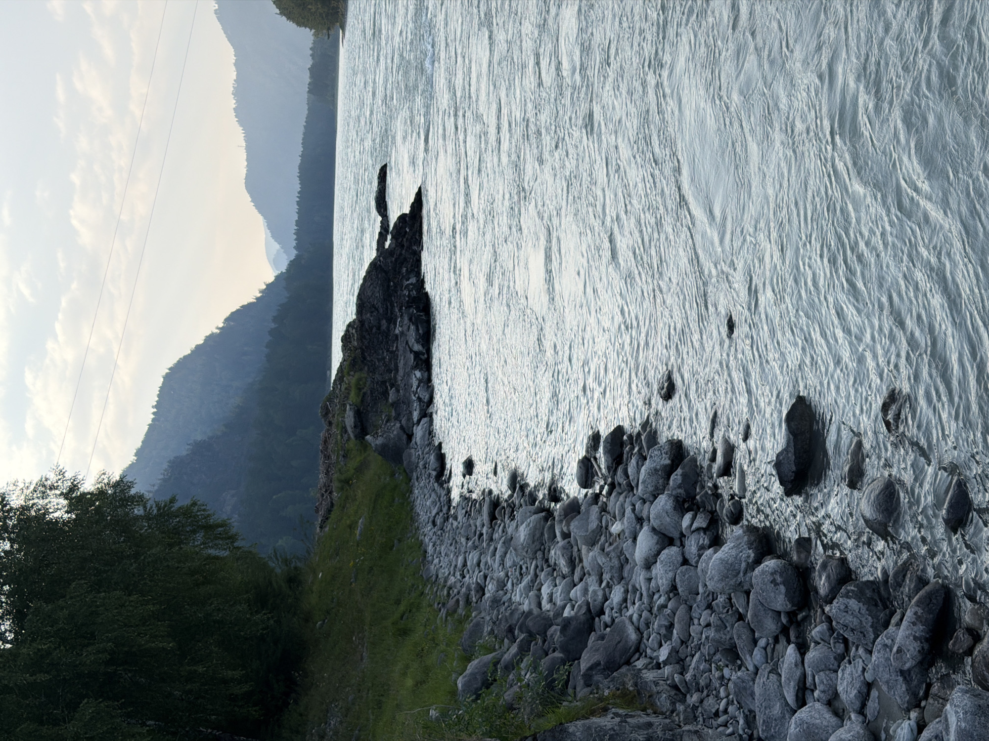 The image depicts a wide, choppy river flowing past a rocky shoreline. On the left, a steep, grassy embankment covered with trees slopes down to the water. A jumble of large, gray stones forms the shoreline, some submerged in the flowing water. In the distance, misty, tree-covered mountains rise, their peaks fading into the overcast sky. The water's surface is textured with ripples and small waves, reflecting the muted light of a cloudy day. Overhead power lines are visible stretching across the sky between the trees and the mountains. The scene conveys a sense of natural, untamed beauty in a remote, mountainous landscape.