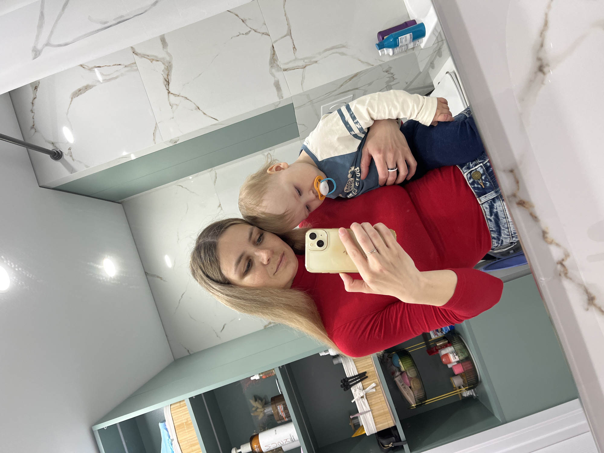 A woman stands in a bathroom with marble-tiled walls and shelves containing toiletries. She is holding a sleeping baby in her arms and taking a selfie with a smartphone. The location is presumed to be in Chaikovsky, Russia.
