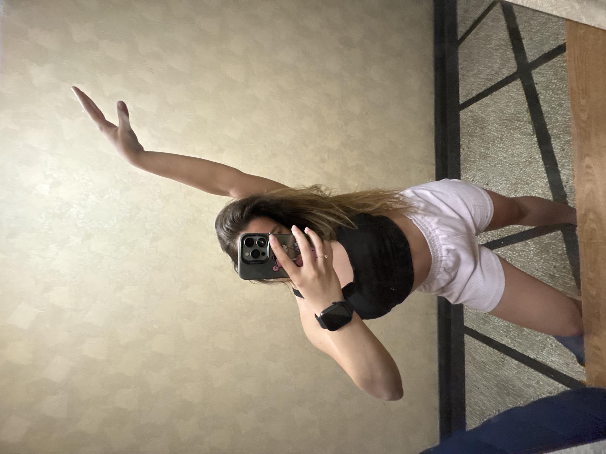 The image depicts a full-body mirror selfie of a young woman taken indoors, in what appears to be a bedroom or personal space in Sofia, Bulgaria. The woman is positioned slightly off-center, with her body angled. She has long, wavy brown hair that cascades over her shoulders and back. She is wearing a black sports bra or crop top and white athletic shorts.
Her left arm is extended upwards and bent at the elbow, with her hand open, as if in a dynamic pose or stretch. Her right arm is bent, holding a dark-colored smartphone directly in front of her face, obscuring it from view. The smartphone has a visible triple-lens camera array and a distinctive case, featuring what appears to be a pink panther-like graphic. A black smartwatch is worn on her left wrist, and a small, simple ring is visible on her right hand's ring finger.
The background reflected behind her is a light-colored wall with a subtle, textured finish. In the lower right portion of the image, the reflection of the floor shows a light-colored surface with a distinct grid or square pattern, alongside a dark blue object (possibly a mat or rug) and a light wooden furniture piece, such as a bed frame or dresser. The lighting is ambient and appears to be from an indoor source. No specific time of day or weather conditions are discernible. There is no legible text visible within the image, beyond the design on the phone case.