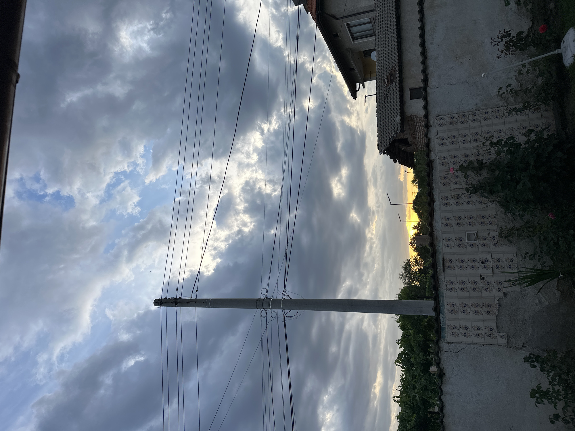 The image captures a view of a cloudy sky with patches of sunlight breaking through, suggesting either dawn or dusk. A tall, concrete utility pole stands prominently in the left foreground, supporting a complex network of overhead wires. To the right, a portion of a building with a tiled roof is visible, showing signs of disrepair with some tiles missing and exposed brickwork. In front of the building is a low wall, partially covered in decorative tiles, and beyond that, a garden area with greenery and flowering plants. The overall scene is one of a suburban or rural environment, with a mix of natural elements and man-made infrastructure.