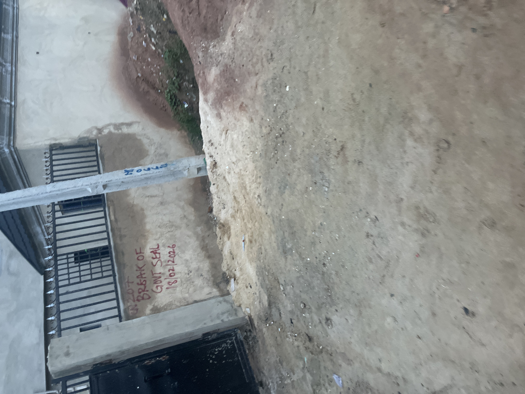 The image depicts a section of a wall in Ikorodu, Nigeria, with graffiti written in red. The graffiti reads "N207 BREAK OF GOVT SEAL 18/02/2026". To the left of the wall is a black metal gate and a concrete pillar. Behind the wall is a metal fence and a building with a tiled roof. To the right of the wall, there is a pile of dirt and some vegetation. A concrete pole with some blue markings is also visible in the foreground, to the left of the graffiti. The ground in front of the wall is unpaved and appears to be dirt and gravel. The scene appears to be an outdoor, public space.