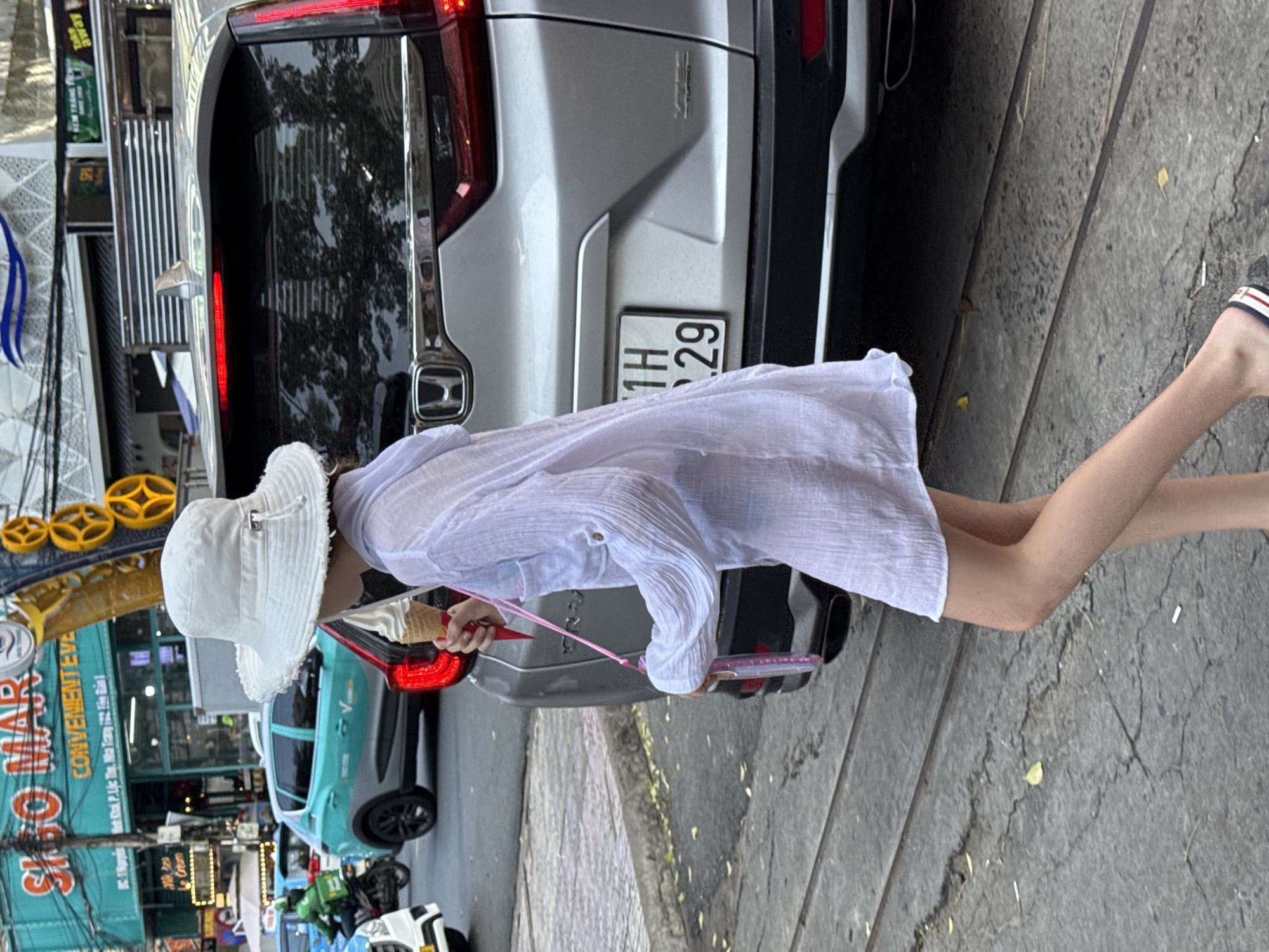 An outdoor, eye-level, full-shot image captures a young person walking on a cracked pavement in Nha Trang Ward, Vietnam. The individual, viewed from a slight rear-right perspective, wears a white wide-brimmed bucket hat with a frayed edge, a sheer white long-sleeved tunic-style top/dress, and striped open-toed sandals. They hold a white soft-serve ice cream cone in a red/pink paper holder in their right hand and a pink-strapped phone case or small bag in their left hand, slung over their shoulder. The person is walking alongside a silver fifth-generation Honda CR-V SUV, which displays "VTEC TURBO" badging and a partially visible Vietnamese license plate reading "1H.29". The background reveals an urban street with other vehicles, including a teal VinFast VF e34 electric car and a green Grab delivery motorcycle with a rider. Buildings line the street, featuring Vietnamese signage such as "SIÊU THỊ CONVENIENT EVE" on a teal structure and "KEM TRANG" (Ice Cream). Decorative yellow circular elements are visible on a nearby pole. The ground consists of cracked concrete or asphalt with parallel grooves, possibly old tram tracks, and scattered leaves. The scene is illuminated by daylight, suggesting a daytime setting.
