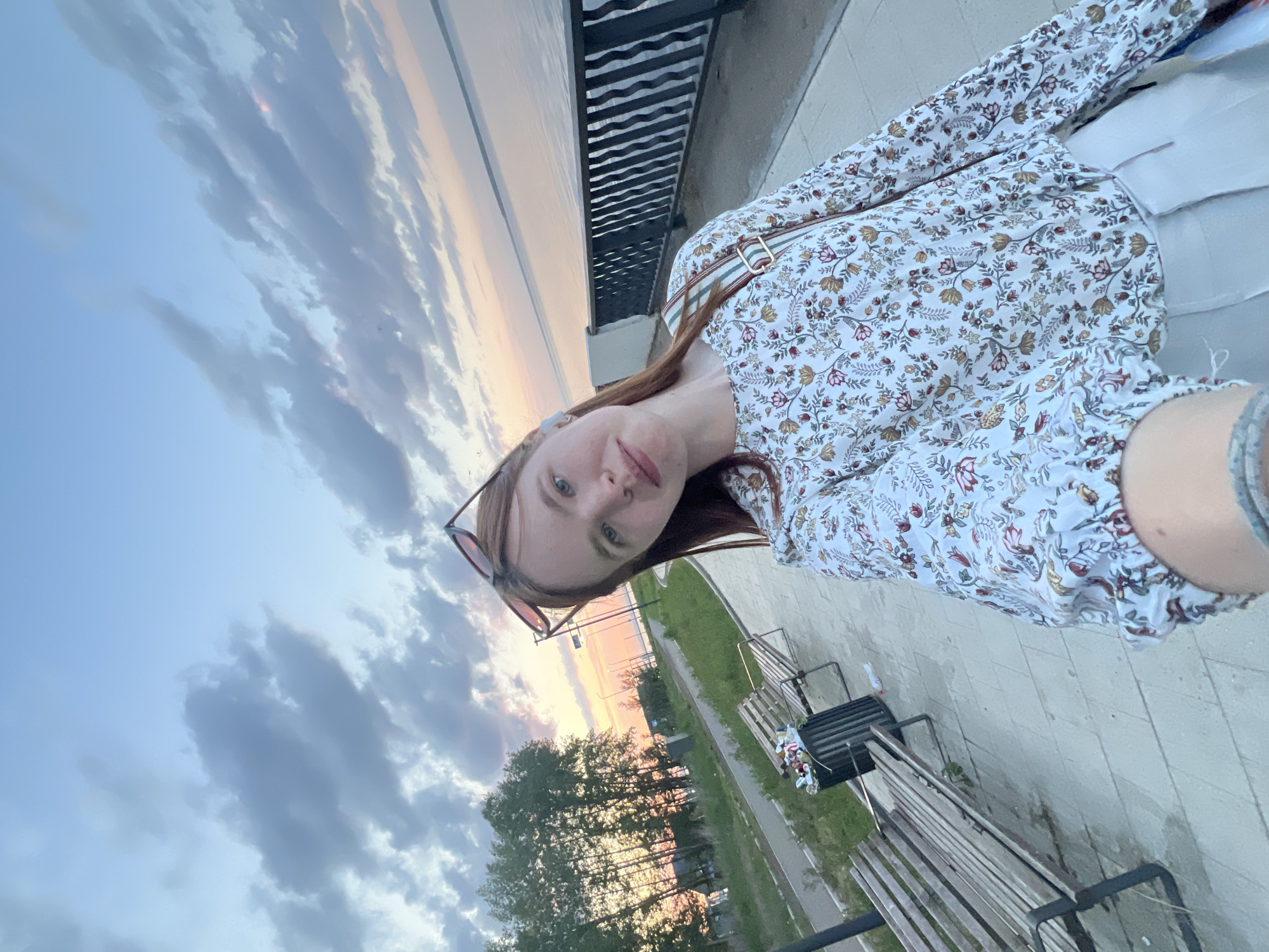 A woman is taking a selfie in the city of Chaikovsky, Russia. The background shows a body of water with a metal railing along the edge of a walkway or bridge. Below the walkway is a green space with trees and benches. The sky has a sunset with pink and orange hues. The woman is wearing a floral patterned blouse and sunglasses are perched on her head.