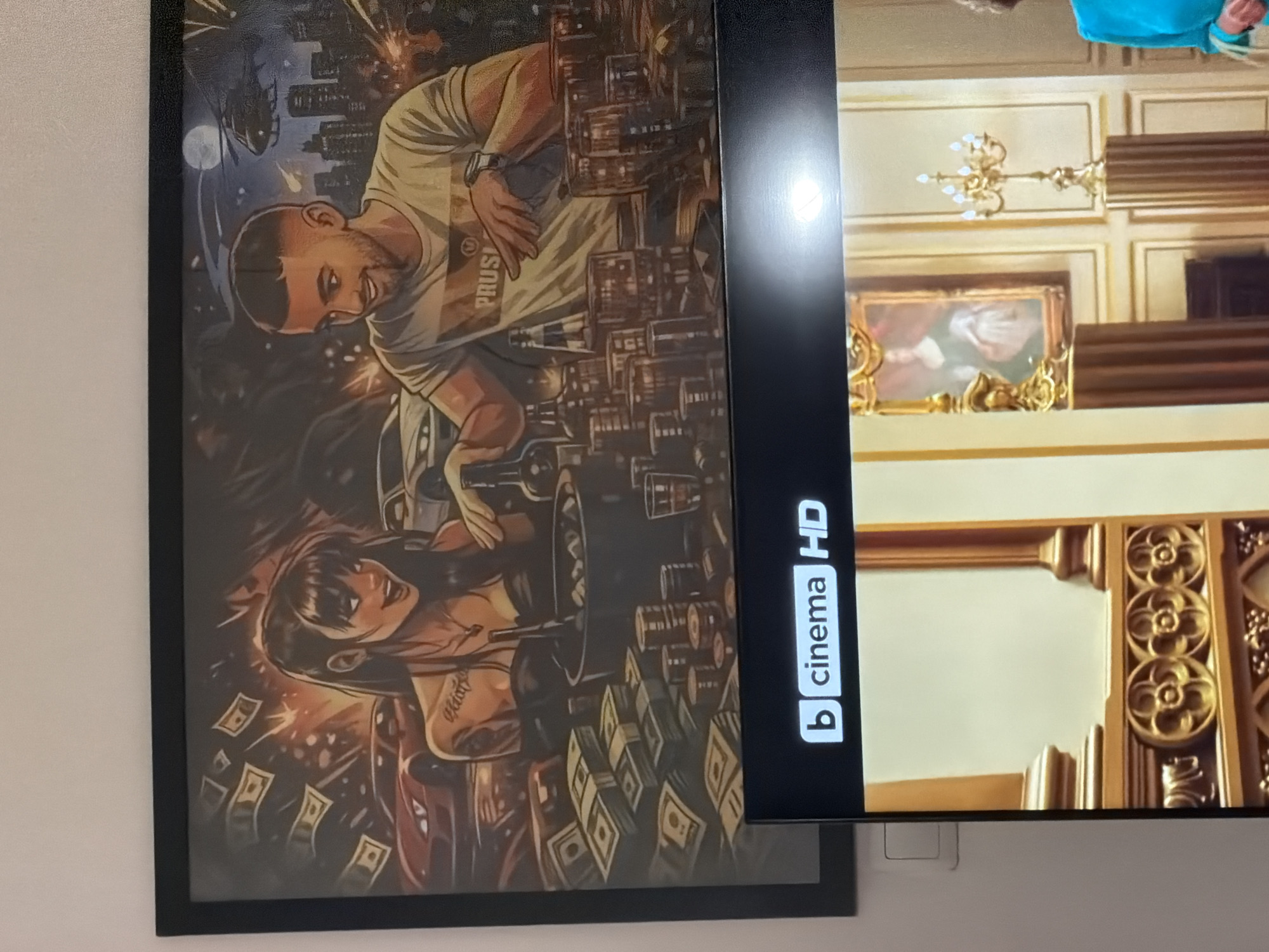 The image displays a framed poster mounted on a wall, positioned to the left of a modern, flat-screen television. The photograph itself is oriented in portrait mode, showing the items rotated 90 degrees clockwise from their typical viewing position.
**Poster Details:**
The poster, rendered in a stylized, dark-toned illustration, depicts a nocturnal, urban scene. In the foreground are two cartoon-like figures. A man with short hair and a beard wears a light t-shirt with "PRUSI" visible on it and a watch on his left wrist. He is shown amidst numerous bottles and glasses, seemingly interacting with them. Beside him, a dark-haired woman with tattoos on her arm smiles broadly. The background includes a city skyline at night with a visible moon. Scattered dollar bills and stacks of money surround the figures, suggesting a theme of wealth or indulgence.
**Television Details:**
The television screen, partially overlapping the poster on the right, is displaying an indoor scene. This scene features rich, ornate golden moldings and decor, including a classic multi-light chandelier and a framed painting of a figure, possibly a woman, in historical attire. A person wearing a blue garment is partially visible at the top edge of the screen.
**Visible Text:**
On the lower-left side of the television screen, a white logo reads "b cinema HD".
The setting is an indoor space, indicated by the wall and ambient lighting.