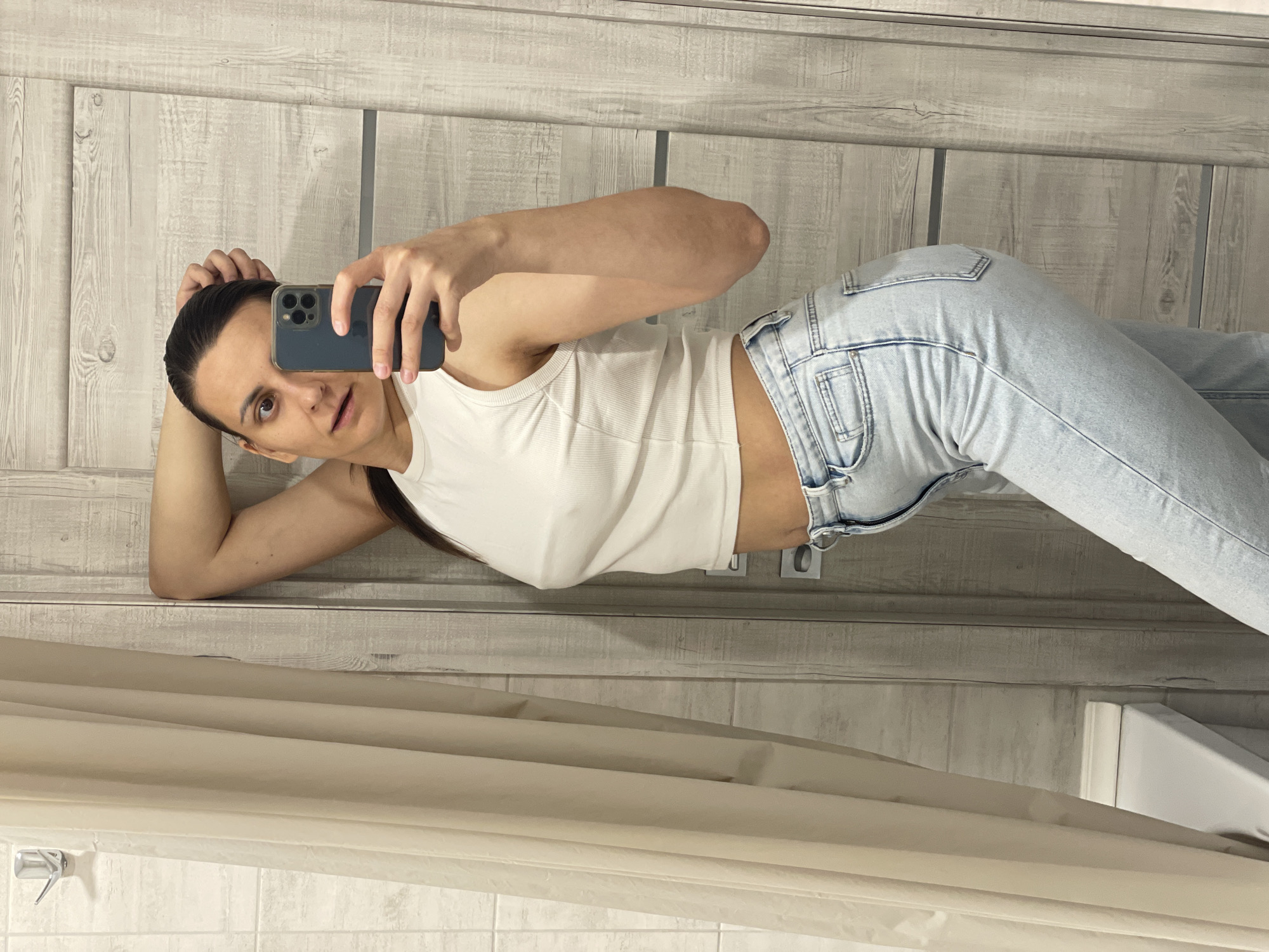 The image captures a woman in what appears to be a bathroom. She is holding a smartphone, presumably taking a selfie. She is wearing a white tank top and light blue jeans. She appears to be positioned against a door or wall with a light wood grain pattern. A shower curtain and part of a bathtub are visible in the lower portion of the image. The scene is not specific to any known city.