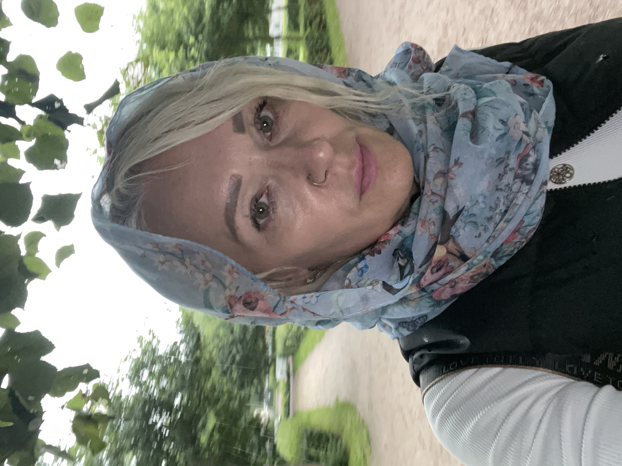 A medium shot selfie shows a woman with blonde hair and green eyes wearing a light blue floral scarf draped over her head and shoulders. She has a nose ring and is wearing light pink lipstick. She is also wearing a dark vest over a white ribbed shirt. The background is blurred, showing a park path with trees and green foliage, suggesting an outdoor setting. The lighting is dim, and there appears to be light rain falling. The scene is likely in Kronstadt, Russia, given the context provided.