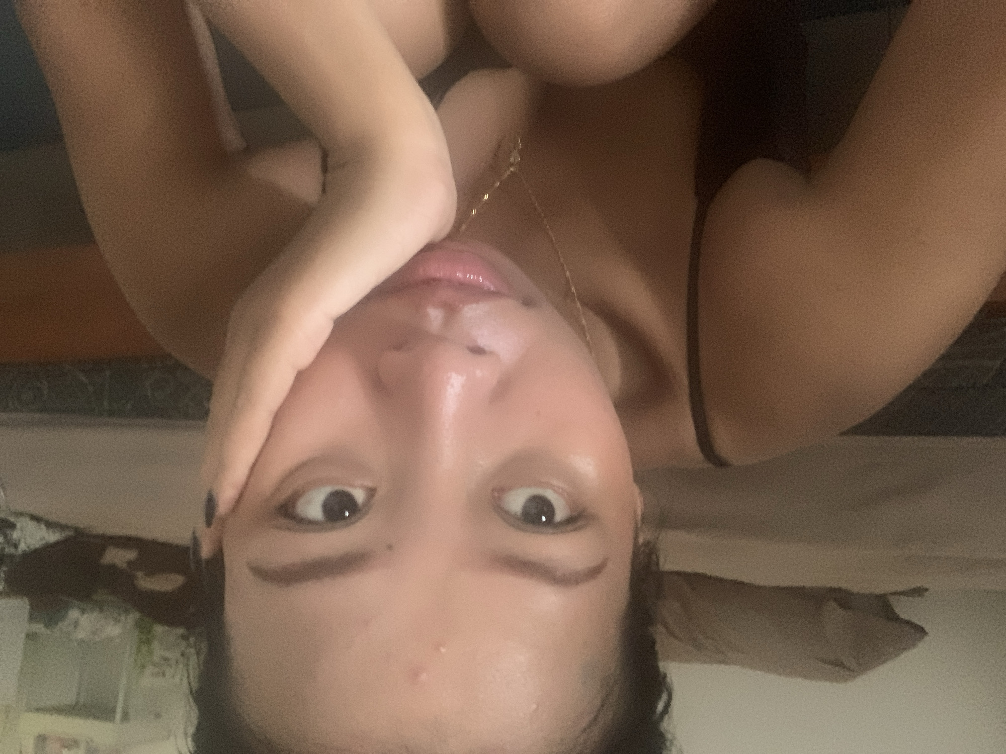 An upside-down close-up portrait of a person with fair skin, dark hair, and dark eyes is presented. The person is holding their hand close to their face. A gold necklace is visible. The background includes a pillow, possibly a bed, and some blurred objects, suggesting an indoor setting. There is no information to suggest that this scene occurred in Denpasar, Indonesia.