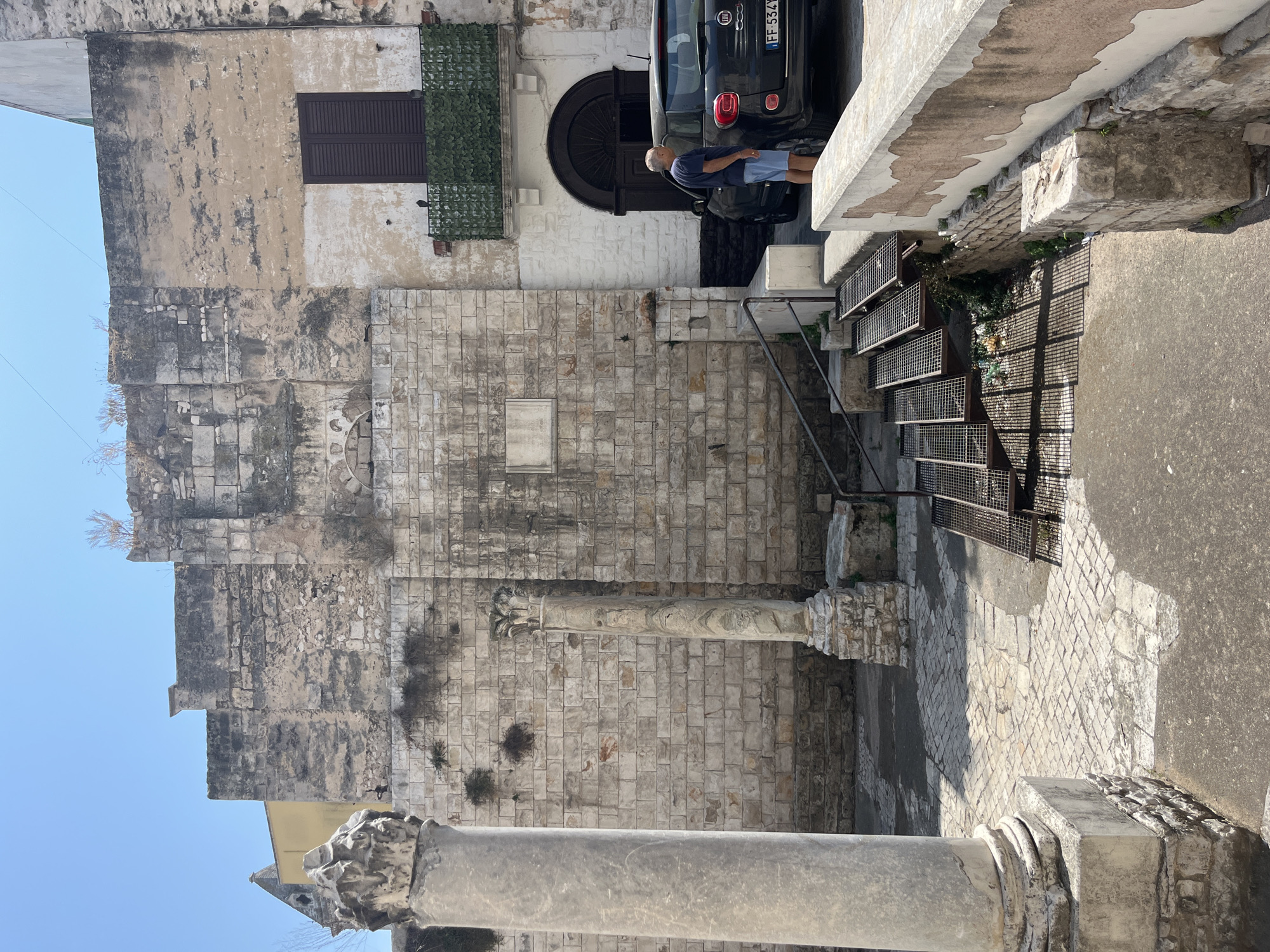 The image depicts an outdoor scene in Bari, Italy, near the Basilica di San Nicola. In the foreground, a man in a blue t-shirt and shorts stands beside a black Fiat 500 car. He is looking at the car. To the left, a large, weathered stone column stands prominently. Behind the column and the man, a stone wall forms the backdrop, with a doorway and a window with dark shutters visible on the right side of the wall. Metal stairs with a grating surface lead up to the doorway. The ground is paved with stones, and shadows are cast by the architectural elements. The overall impression is of an old, historical urban environment.