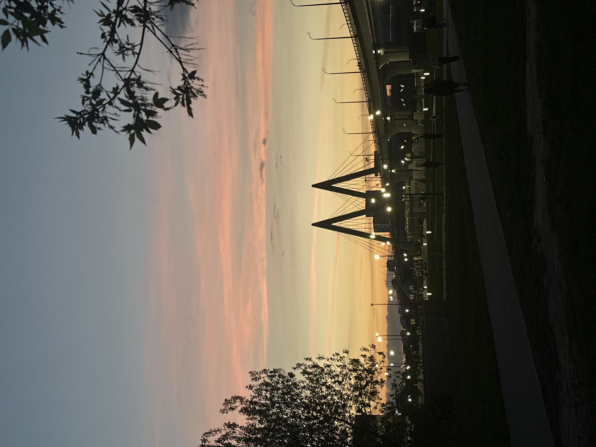 The image shows a sunset over the city of Kazan, Russia. A distinctive cable-stayed bridge with two A-frame pylons is visible. Many street lights line the walkway and edge of the frame. The sky transitions from a pale blue to a peach and orange gradient. Silhouetted trees occupy the foreground. Silhouetted figures of people are visible on the walkway.