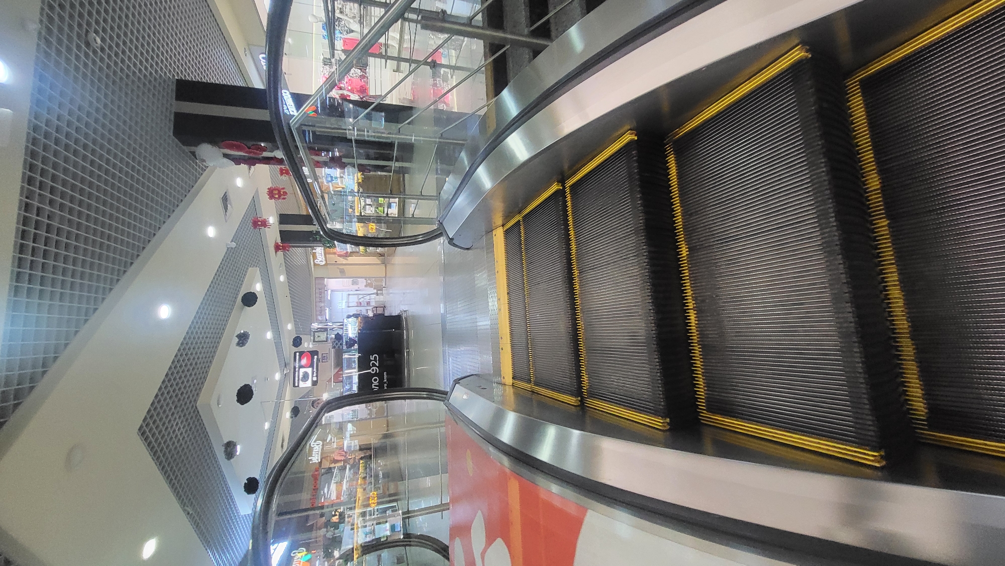 This image captures a view of a shopping mall interior, specifically focusing on an escalator. The perspective is looking down the escalator, with multiple steps visible in the foreground. The escalator treads are dark gray with bright yellow trim along the edges, and they are angled downwards. The handrail appears to be made of a metallic material, possibly stainless steel, and is curved as it follows the path of the escalator.
To the left of the escalator, there is a glass railing that separates it from the rest of the mall. Beyond the railing, the interior of the mall is visible, including a patterned ceiling with square tiles, recessed lighting, and decorative elements. Some shops or display areas are visible in the background, with merchandise and signage. There's a black kiosk or information desk with some signage on it.
The scene suggests a typical day in a shopping mall. There are no people prominently featured in the foreground, but some figures might be present in the distant background or on other levels of the mall. The lighting appears to be artificial, indicating that it could be any time of day. The overall impression is one of a modern, commercial space designed for shoppers.
Visible text includes "uno 925" on a black kiosk. Other text is too small or blurry to decipher.