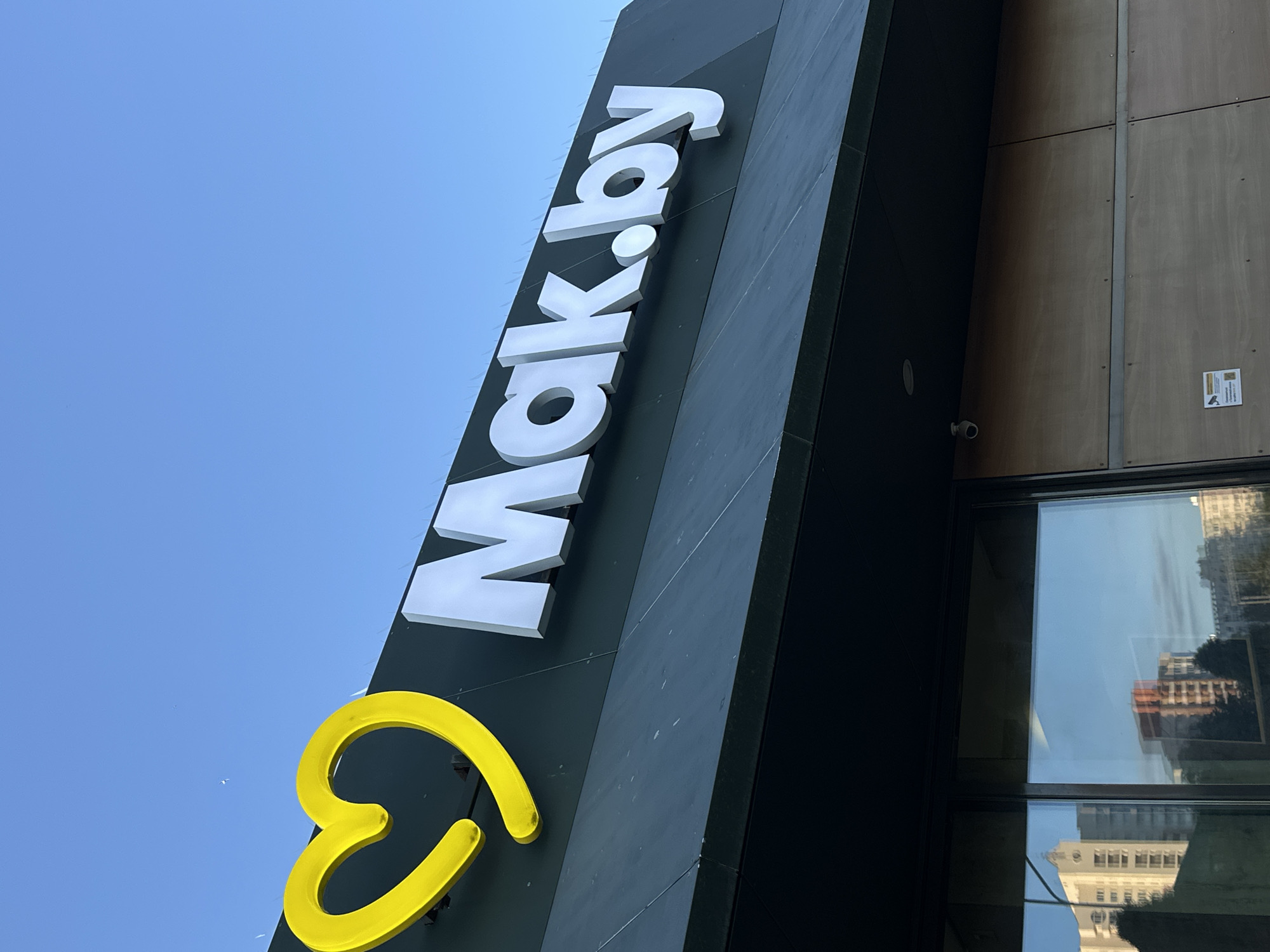The image shows a close-up view of the exterior signage of a fast-food establishment, identified by the prominent "Mak.by" logo. A large, bright yellow heart symbol is positioned to the left of the white, three-dimensional lettering. The sign is mounted on a dark, slanted surface that appears to be the facade of the building. The sky above is clear and blue. Below the sign, the upper part of a glass storefront is visible, reflecting buildings and trees from across the street. The architectural style of the reflected buildings suggests an urban environment, consistent with a location in Minsk, Belarus, where "Mak.by" is the operating name for McDonald's. A small security camera is visible mounted on the building above the storefront.