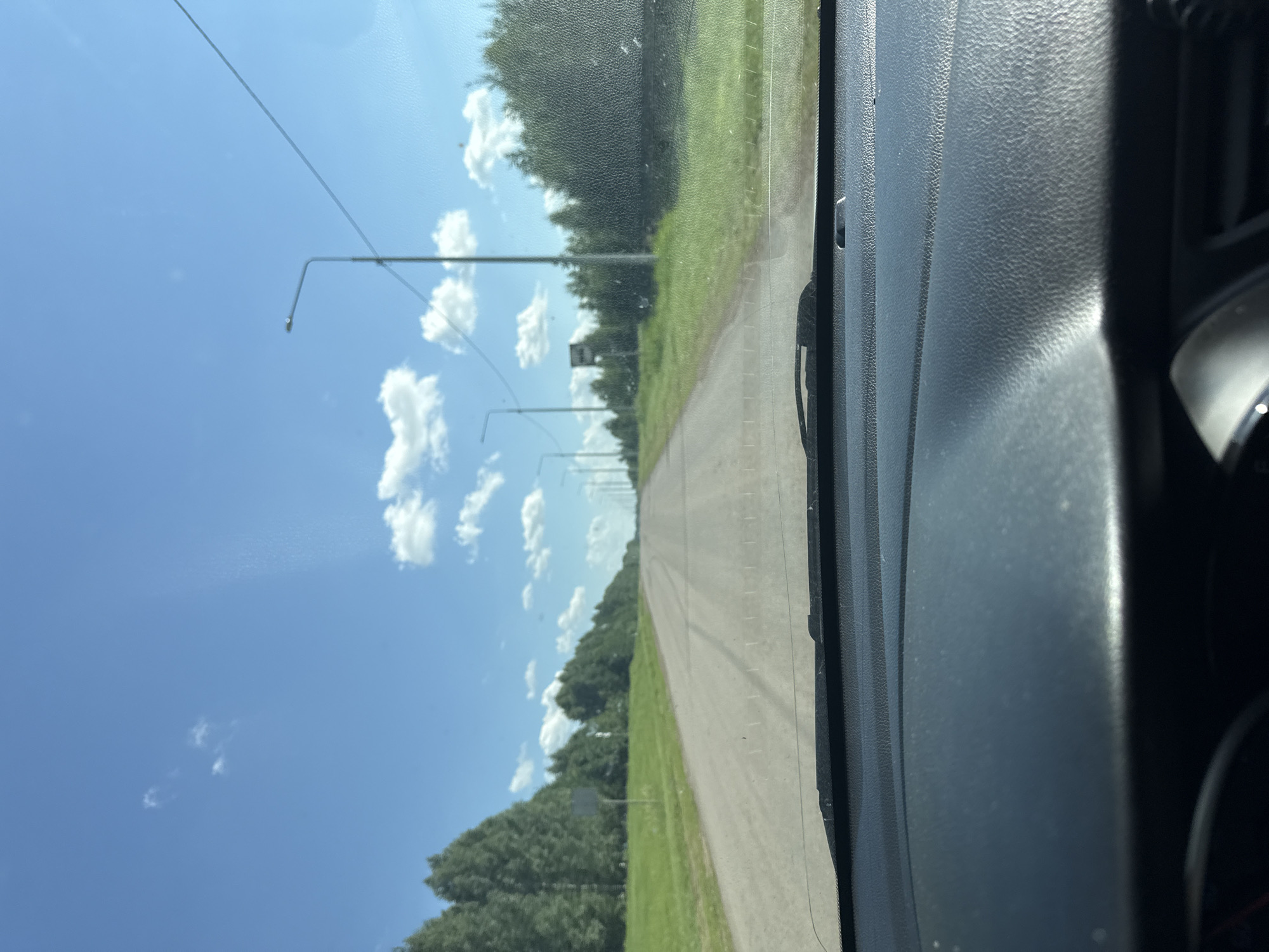 The image shows a view from inside a car looking out at a straight, paved road lined with trees on both sides. The sky is blue with scattered white clouds. There are streetlights visible along the side of the road. The dashboard of the car is visible in the foreground. The scene appears to be in a rural or suburban area. This image is consistent with the landscape of Verkhneyarkeyevo, Russia, a town located in the Republic of Bashkortostan, known for its natural beauty and agricultural surroundings.