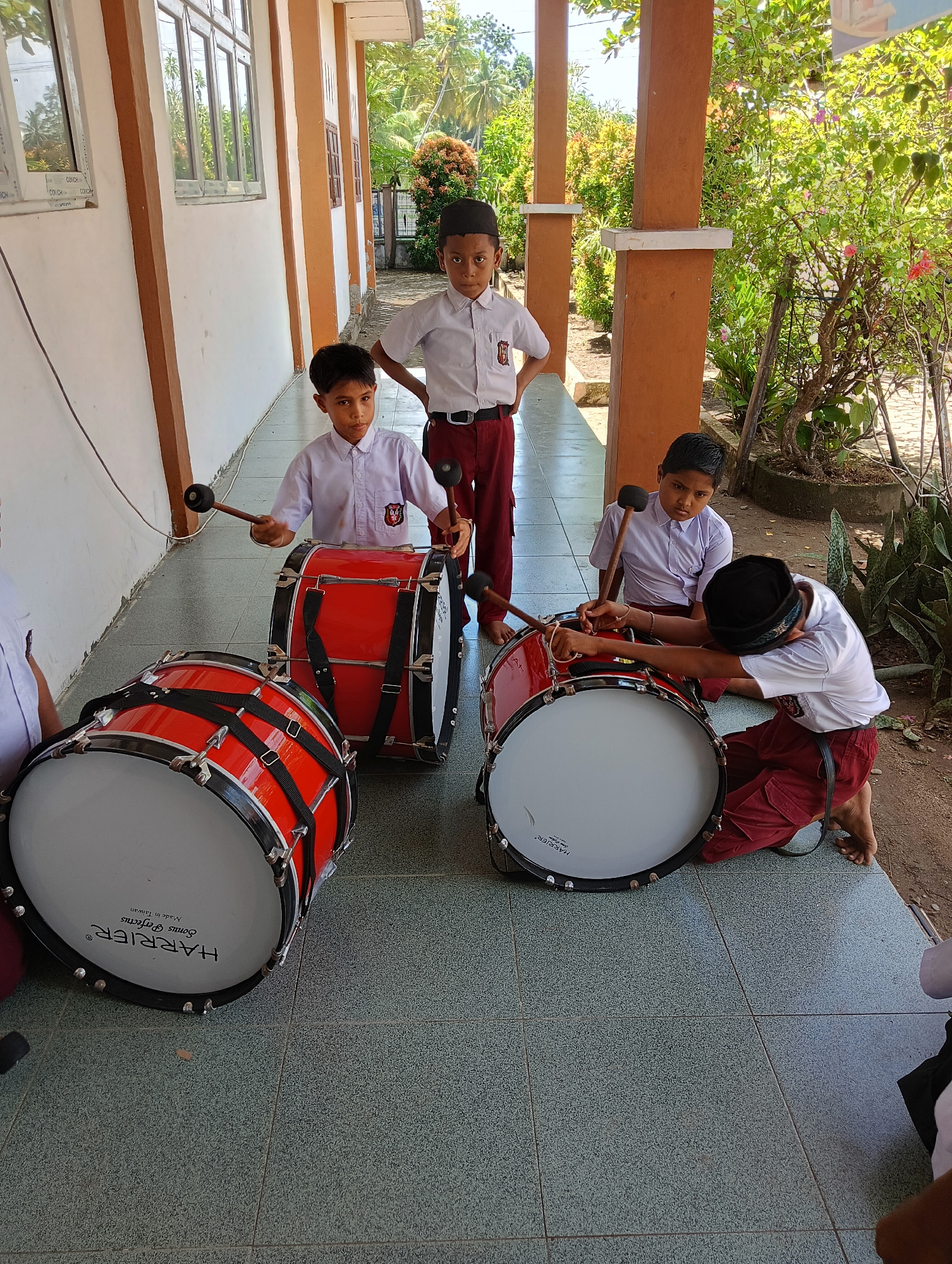 In Baratpayaitek, Indonesia, four young boys in white shirts and maroon pants practice with large red bass drums outside a building. The building has an orange and white exterior, with a tiled walkway in front. Two of the boys are standing behind the drums, holding mallets, while one boy is kneeling beside a drum, and another is partially visible on the left. Lush green foliage and trees are visible in the background.