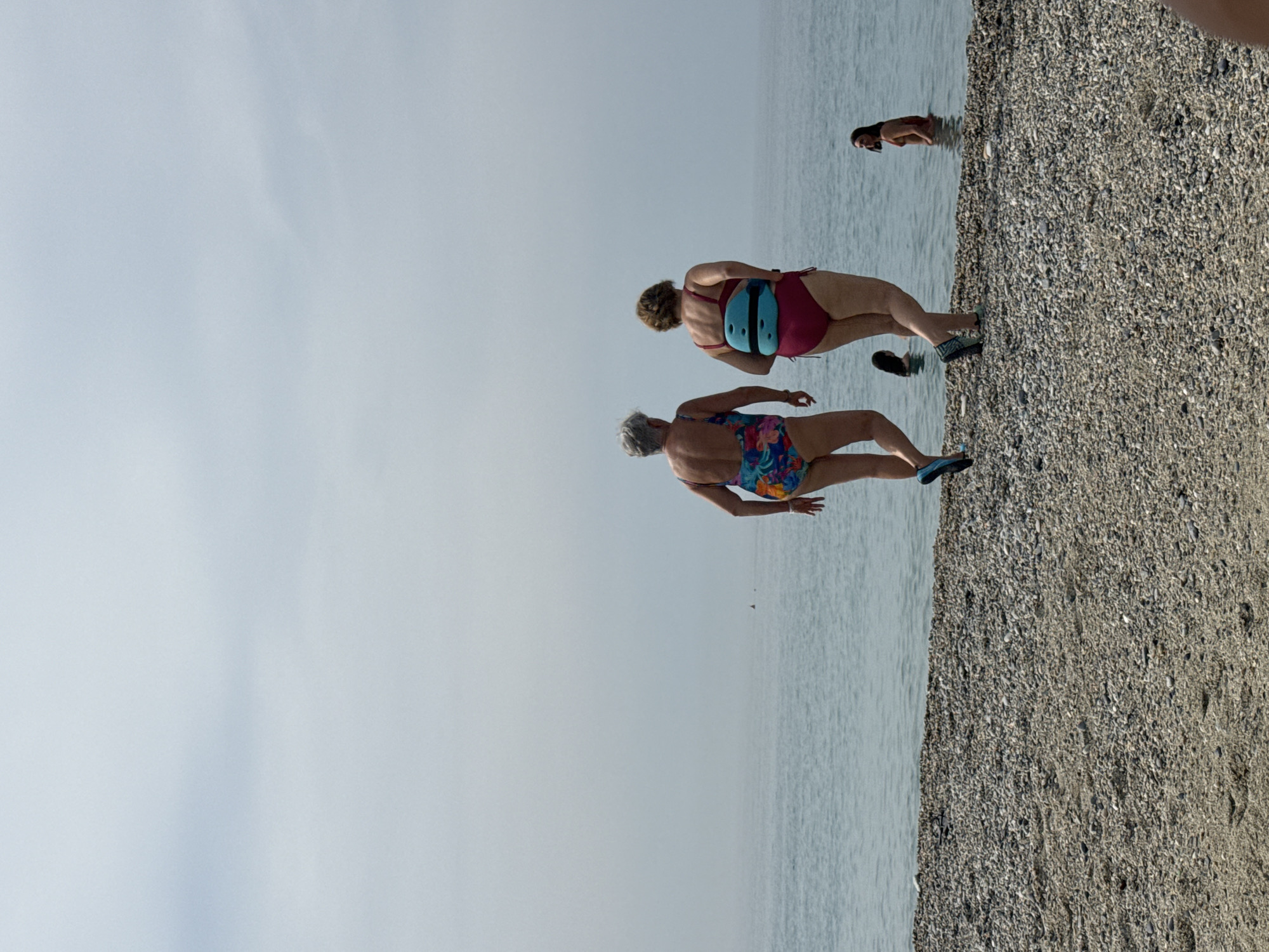 Two women in swimsuits walk on a pebble beach towards the sea. The woman on the left wears a floral-patterned swimsuit. The woman on the right wears a solid red swimsuit and a teal floatation device around her waist. In the water behind them, two other people are visible. The sky is overcast. This scene is located on the beach in Finale Ligure, Italy.