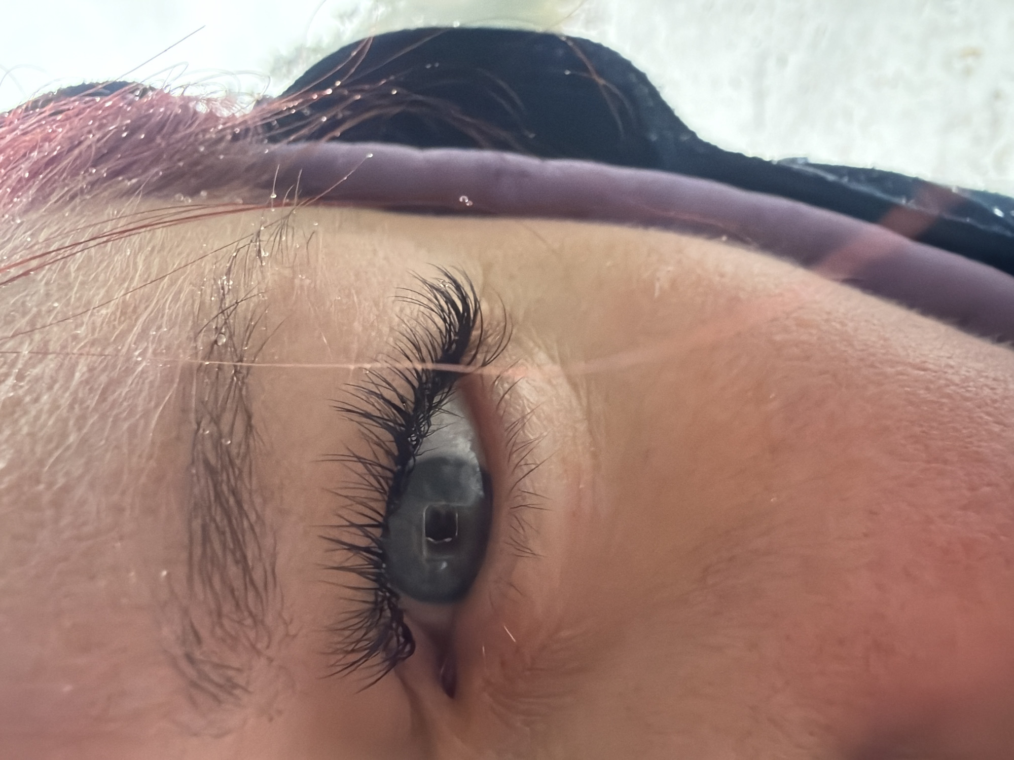Close-up of a person's eye with long, dark eyelashes, lying on their back. Light gray or blue iris. The person has pink-tinted hair. Some water droplets are visible on the hair and skin. The person is wearing a black garment with a purple pillow or cushion under their head. There is no indication of the location being Chaikovsky, Russia, or any specific city.