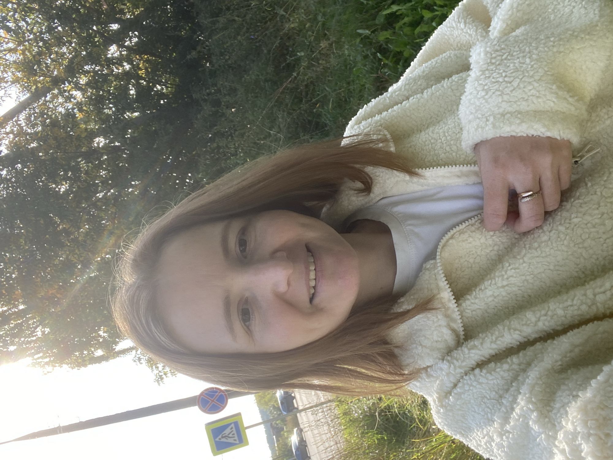A young woman with shoulder-length auburn hair smiles at the camera, holding her left hand to her chest. She wears a cream-colored, fuzzy zip-up jacket over a white t-shirt and two rings on her left ring finger. The background features a street with cars driving by, a pedestrian crossing sign, and a utility pole with the sun shining through trees. The setting appears to be a street in the city of Chaikovsky, Russia.