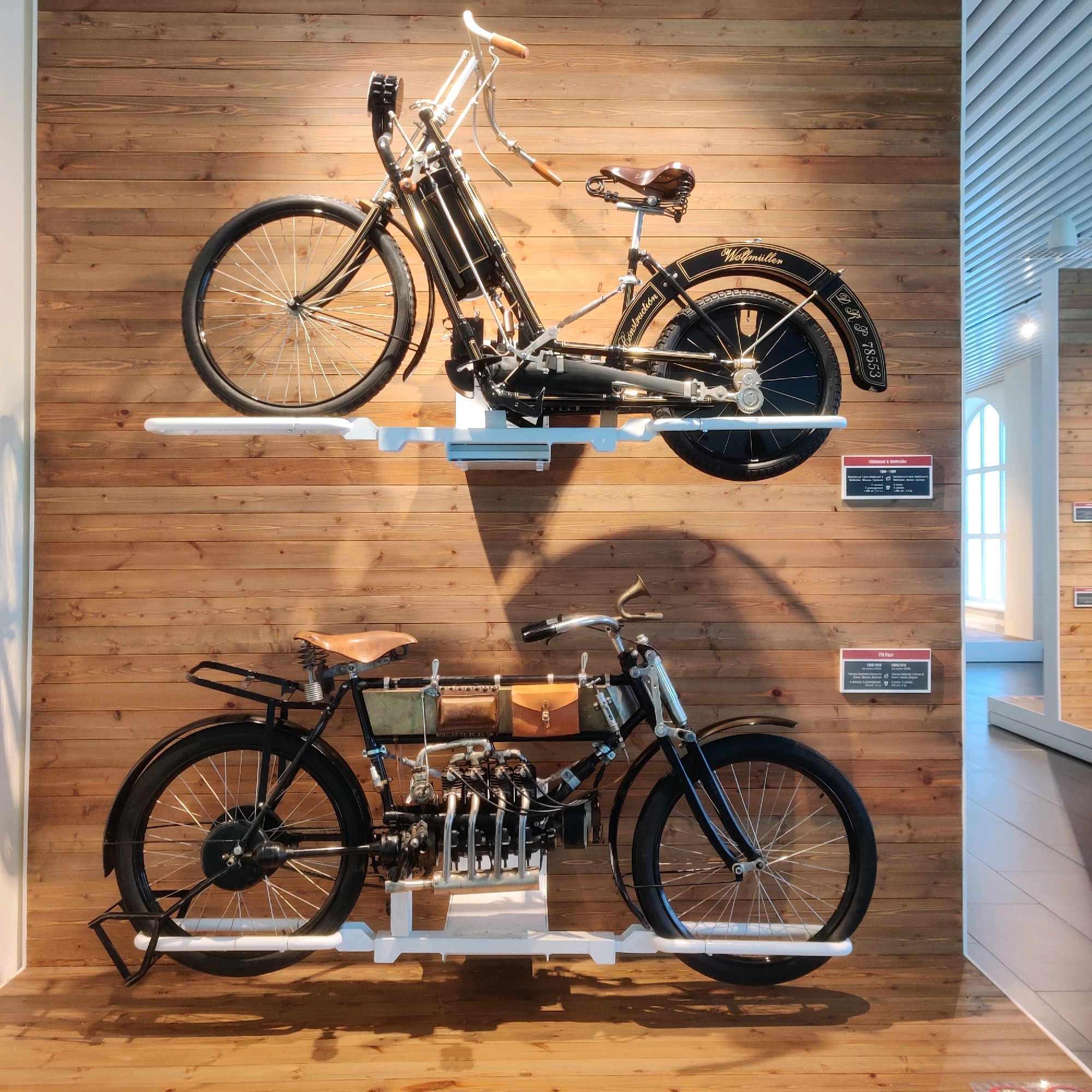 The image displays two antique motorcycles mounted on a wooden wall in a museum. The motorcycles are positioned one above the other, each on a white display stand. The upper motorcycle is black with gold trim and features the name "Wolfmüller" on its rear fender. The lower motorcycle is also black and appears to have a more complex engine with multiple cylinders. Both motorcycles have brown leather seats and spoked wheels. The wall is made of horizontal wooden planks. In the background, a brightly lit hallway with white ceilings and windows is visible, suggesting an indoor exhibition space. The overall scene is static and well-lit, highlighting the historical artifacts. The location is the "Ural Military History Museum" in Verkhnyaya Pyshma, Russia, specifically the exhibition dedicated to the history of automotive and aviation technology.