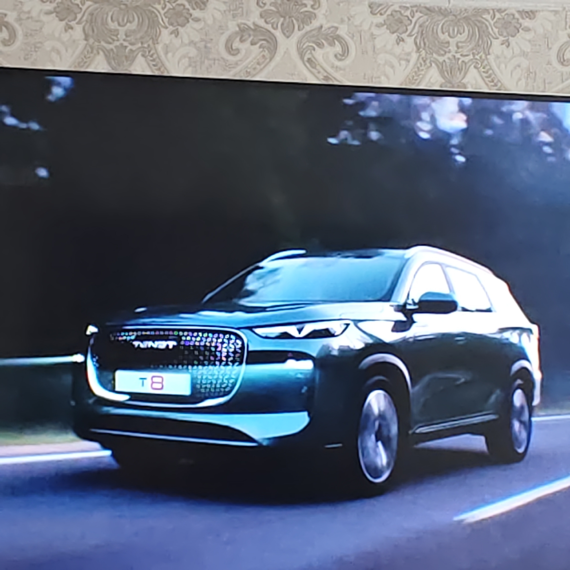 The image shows a dark teal SUV driving on a road. The car has a license plate with "T8" in pink and white. The car's grille is illuminated with colorful lights. The background is blurred, suggesting motion, and includes trees and a sky. There are no people visible in the image. The image appears to be a still from a video advertisement or promotional material for the car.