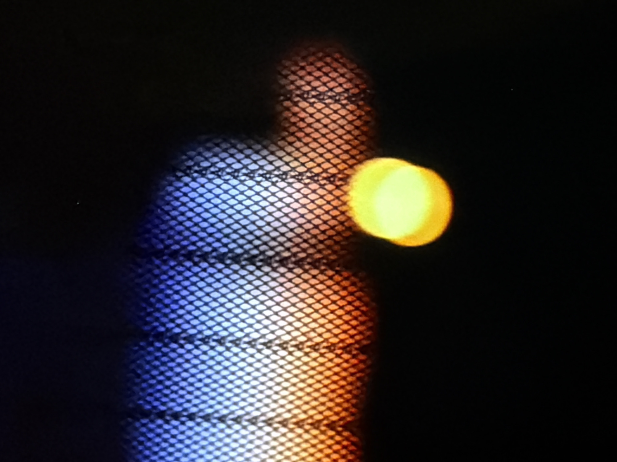 The image is a close-up, low-light photograph of a textured surface, possibly a fence or netting, with a gradient of colors behind it. The top portion of the surface shows a blue hue, transitioning to orange and red at the bottom. In the foreground, a bright, out-of-focus yellow-green circle is visible on the right side of the frame, against a black background. The image appears to be taken at night or in a very dark environment.