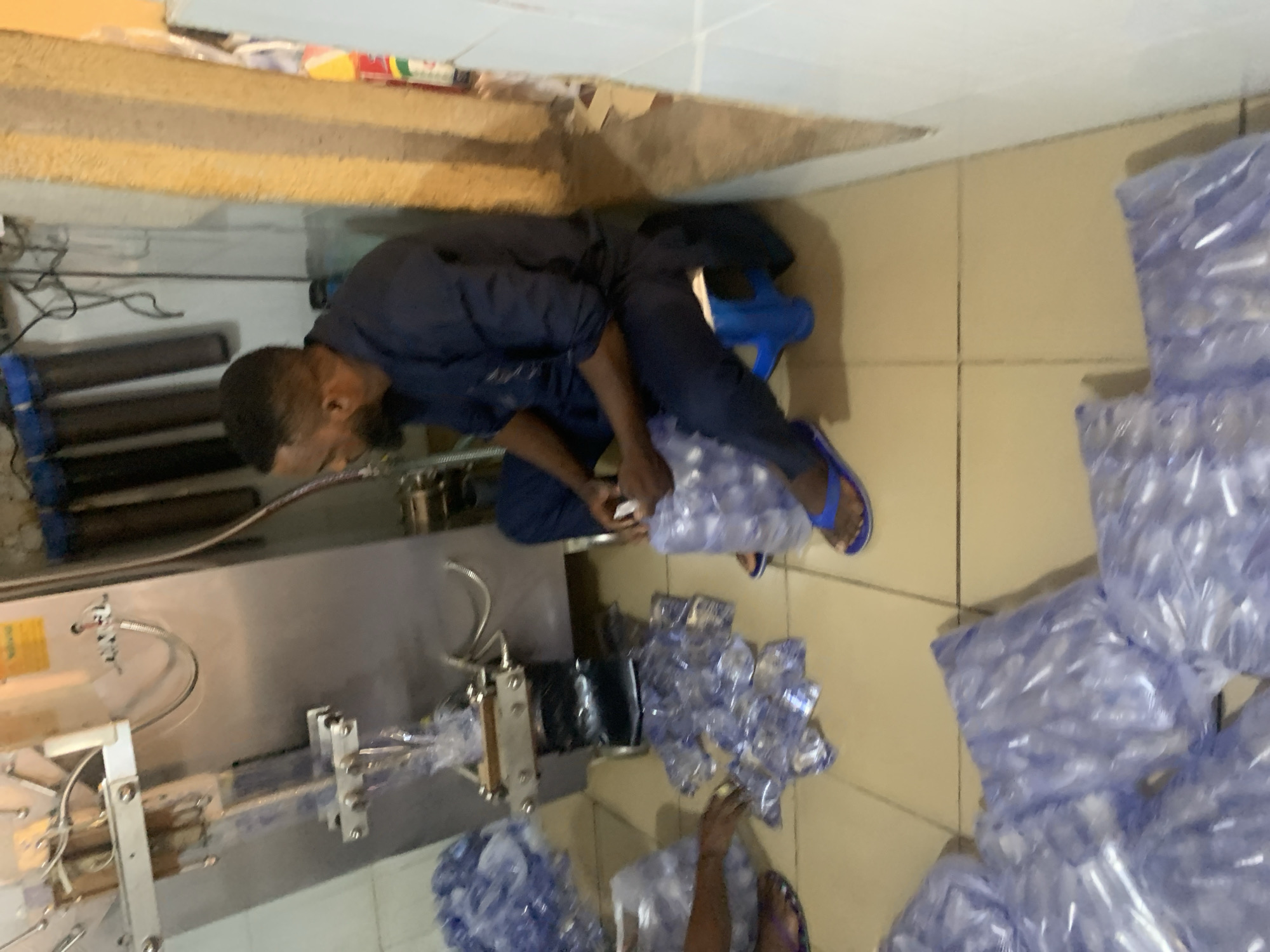 In Rafin Par, Nigeria, a man in a dark blue traditional outfit is seen sitting on a blue stool, facing a water pouch packing machine. He is holding a small, clear plastic pouch, likely a sachet of water, in his hands, appearing to be inspecting or handling it. The machine is a stainless steel unit with a conveyor belt and sealing mechanism. Various stacks of packed water sachets are visible on the floor around him, indicating a production or packaging operation. The floor is tiled in a light beige color, and the environment appears to be an indoor industrial or workshop space, with pipes and other equipment visible in the background. The lighting is somewhat dim.
