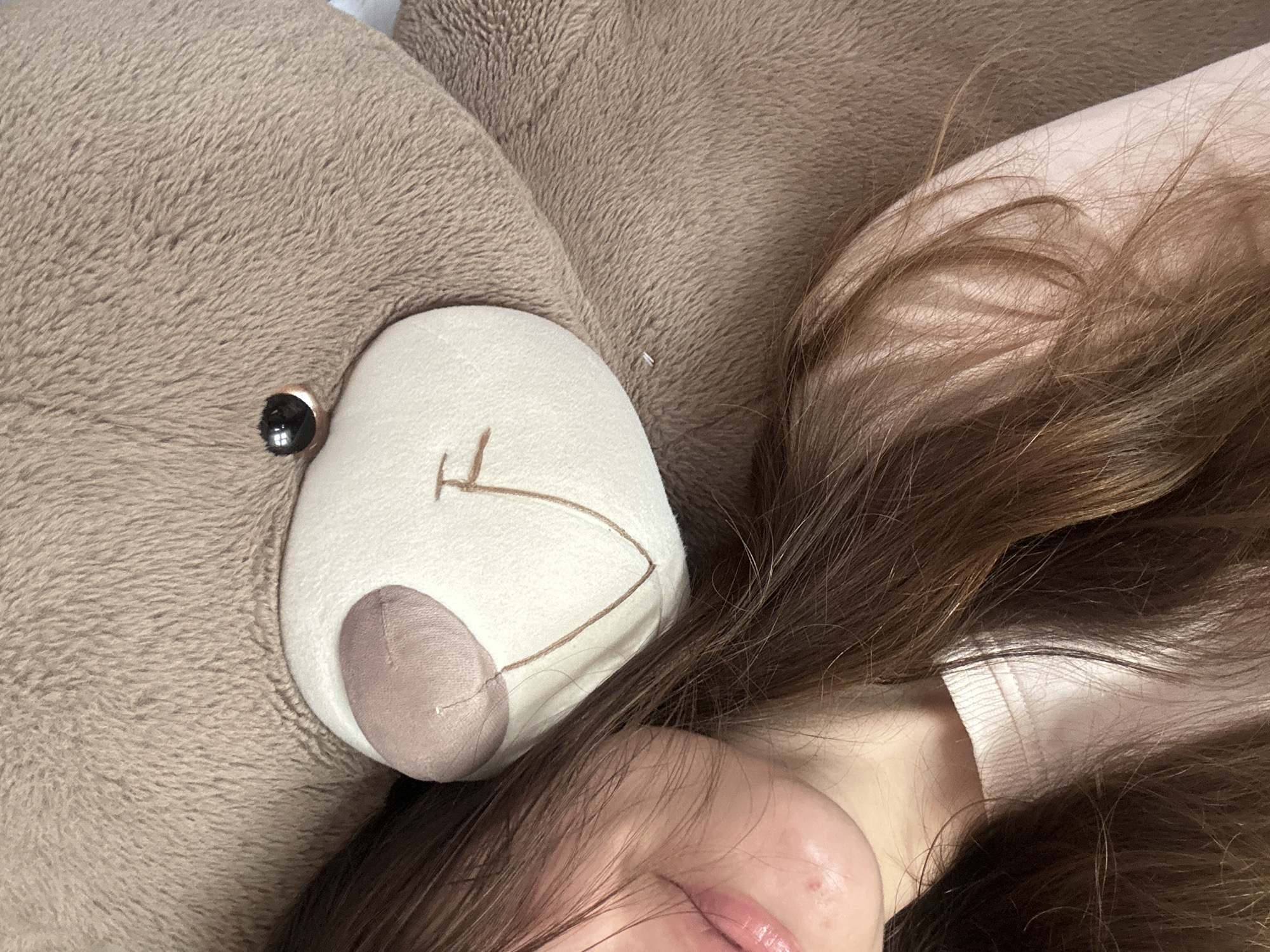 A photograph shows a close-up view of a person lying next to a large, brown stuffed bear. The person's face is partially visible, featuring a portion of their forehead, eye, and lips. The person has brown hair which is splayed out. The stuffed bear is positioned adjacent to the person's face. The bear has a light-colored muzzle and a brown nose. The background is blurred and out of focus. The scene is indoors. The city and state are unknown.