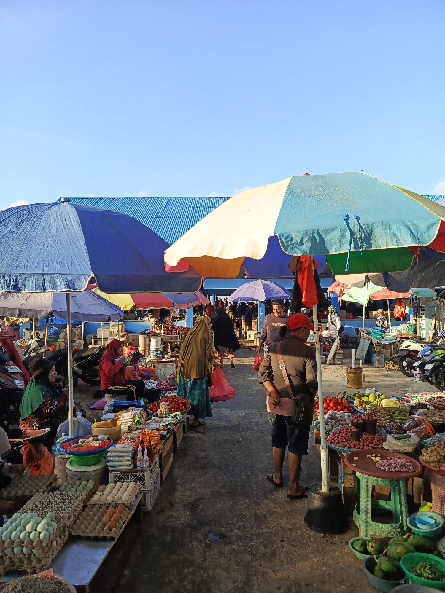 This is a photograph of an outdoor market in Kendari, Indonesia. The market is bustling with activity, featuring numerous stalls selling various goods. The stalls are covered by colorful umbrellas and awnings, providing shade from the sun. In the foreground, several people are browsing and shopping. One person, wearing a red cap, is walking away from the camera. To the left, a woman in a green headscarf is seated behind a display of eggs. Another woman, wearing a brown headscarf and a long skirt, stands near a stall laden with vegetables and other produce. Behind the stalls, a blue-roofed building is visible, suggesting the market is located near a larger structure. The ground is paved with concrete, and motorcycles are parked to the side, indicating transportation is a common mode of arrival for vendors and shoppers. The sky above is clear and blue. The scene depicts a vibrant and active marketplace, typical of many urban centers in Indonesia.