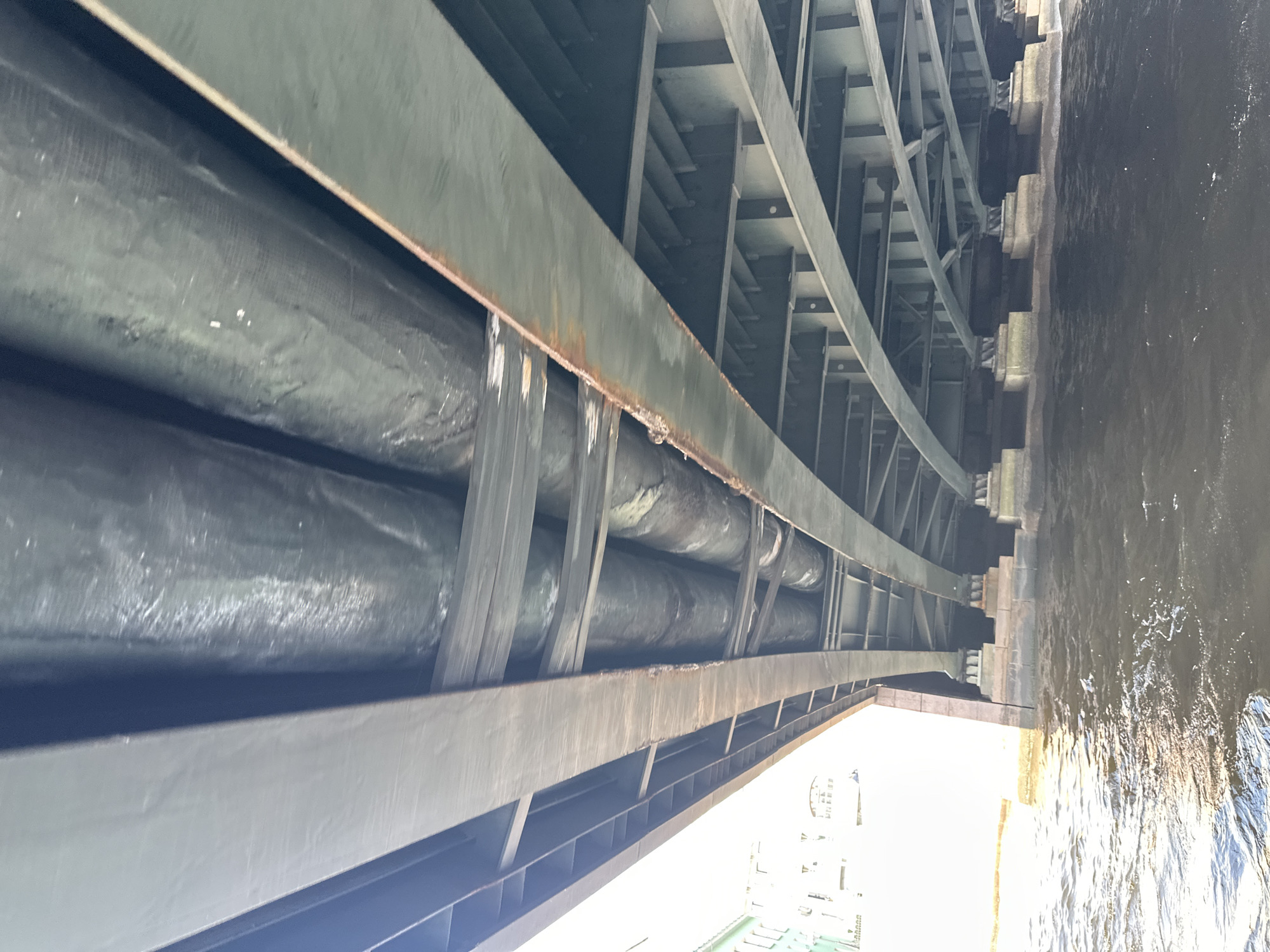 The image captures the underside of a bridge in Saint Petersburg, Russia, revealing a network of steel beams and supports. Two large, cylindrical pipes run parallel to the bridge's curve, secured beneath the structure. The image suggests the pipes might be utility lines. The bridge spans a body of water, visible as dark ripples on the right side of the frame. The overall lighting is bright, but the underside of the bridge is shaded, creating a contrast. The scene is devoid of people. A partial view of buildings and possibly a boat is visible through the opening near the edge of the bridge, adding context to the location.