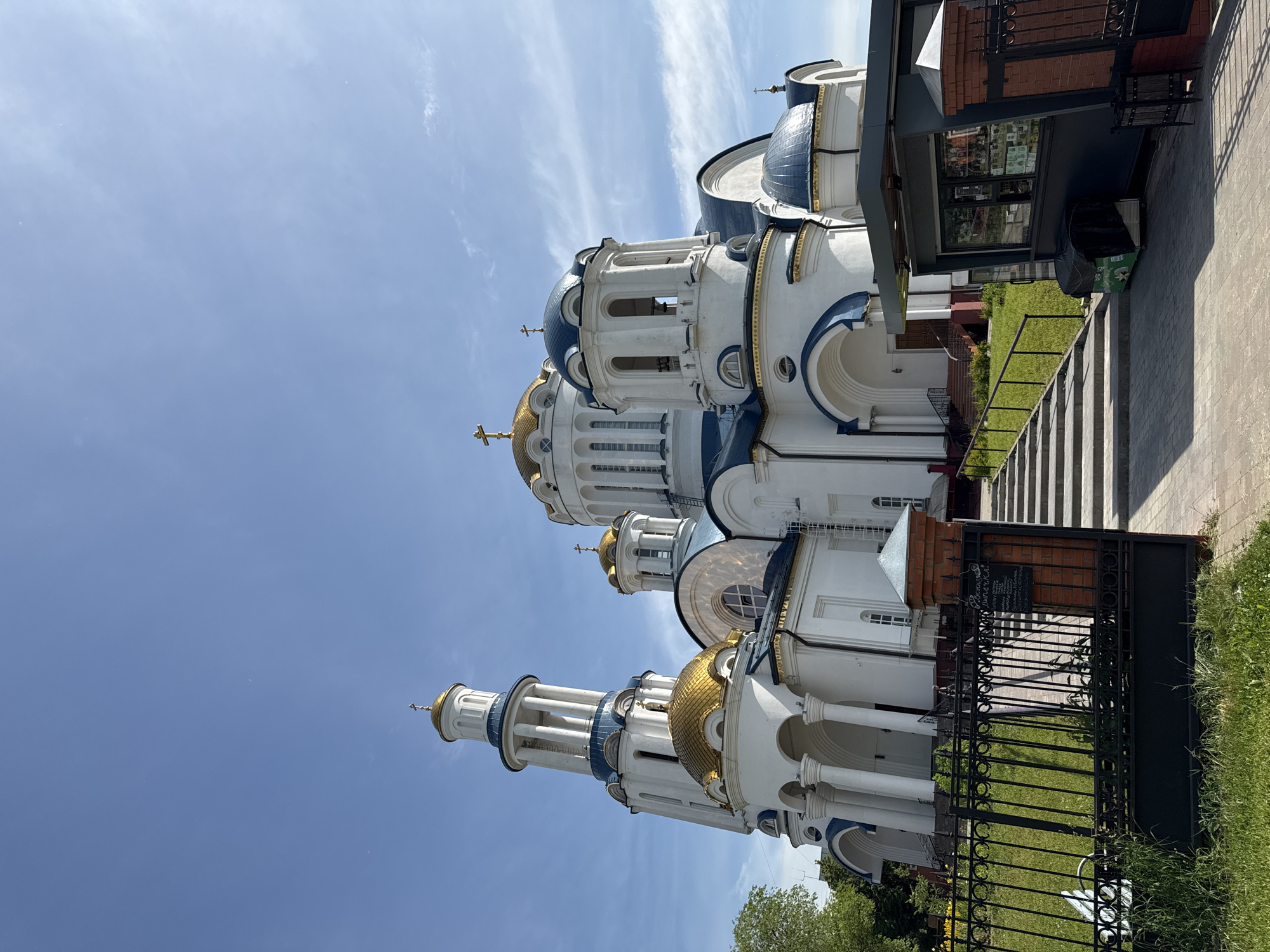 The image shows a large, white Orthodox church with blue and gold domes, set against a clear blue sky with scattered clouds. The church has a tall bell tower on the left and multiple smaller domes and cupolas. There is a black metal fence in the foreground, with green grass and some trees visible to the left. To the right of the fence and grass is a paved pathway leading up to the church entrance, with a small kiosk or shop on the right side of the pathway. The architecture suggests it is a Russian Orthodox church, possibly located in a suburban or park-like setting within Moscow, Russia. The lighting indicates it is daytime. There are no people visible in the image.