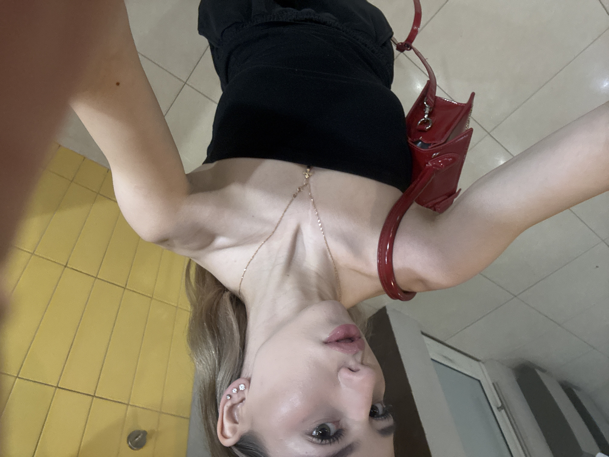 A selfie shot from a high angle depicts a woman wearing a black top and a gold necklace. She has light blonde hair, multiple piercings in her left ear, and noticeable makeup. A red purse hangs from her shoulder. The background is divided into two sections: the left side shows yellow tiled flooring, while the right side displays a white door and wall. The location is not discernible from the image.