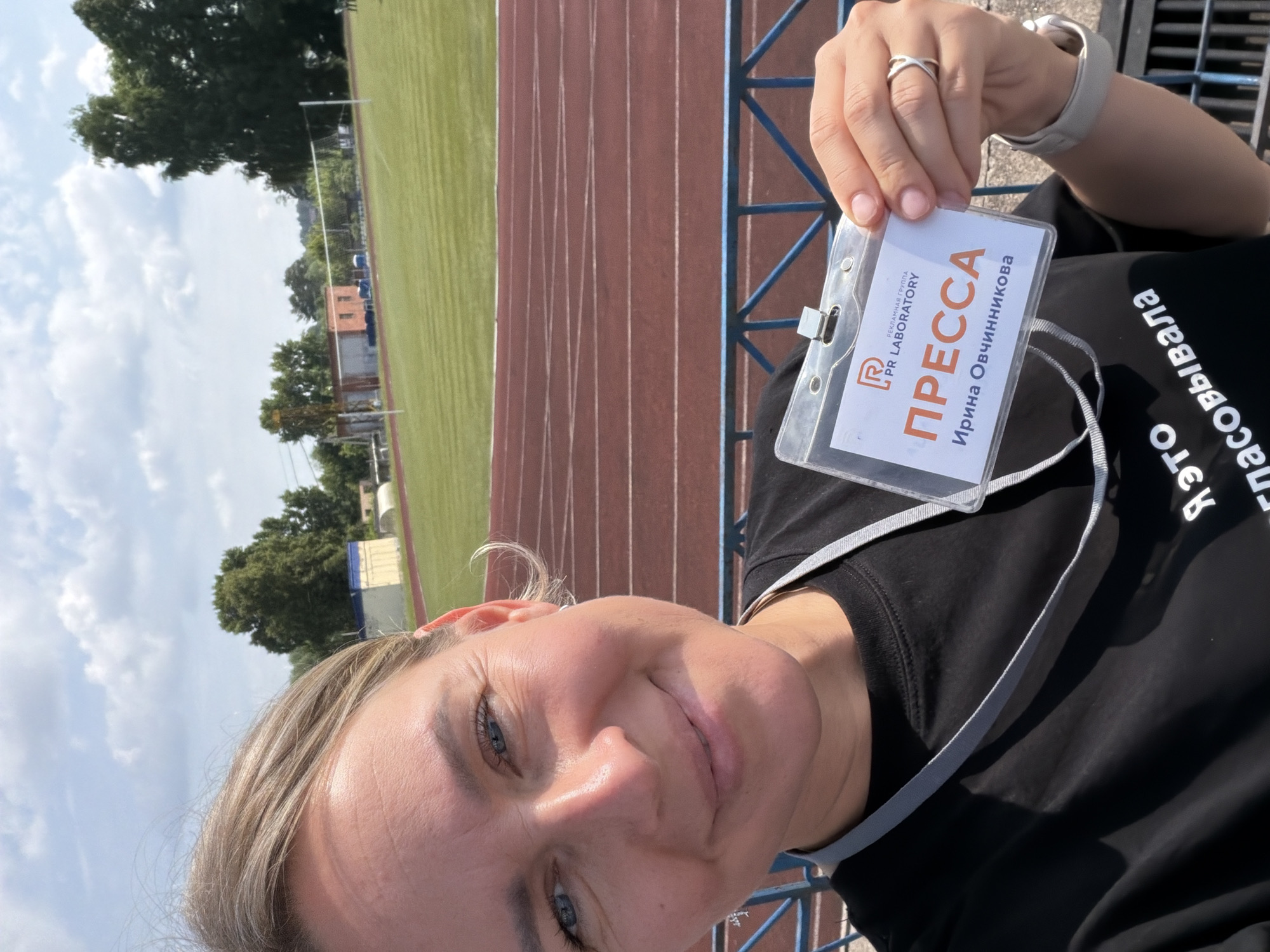 A blonde woman with blue eyes smiles for a selfie at an outdoor athletic stadium. She is wearing a black t-shirt and a grey lanyard with a press pass. The pass has the PR Laboratory logo and the word "ПРЕССА" (Press) in large orange letters, with "Ирина Овчинникова" (Irina Ovchinnikova) written below in smaller text. The stadium features a red running track and a green field. In the background, there are trees and buildings. The woman is wearing a ring on her left hand and a grey fitness tracker on her right wrist. The image appears to be taken on a sunny day with some clouds in the sky. The athletic stadium is located in the city of Chaikovsky, Russia.