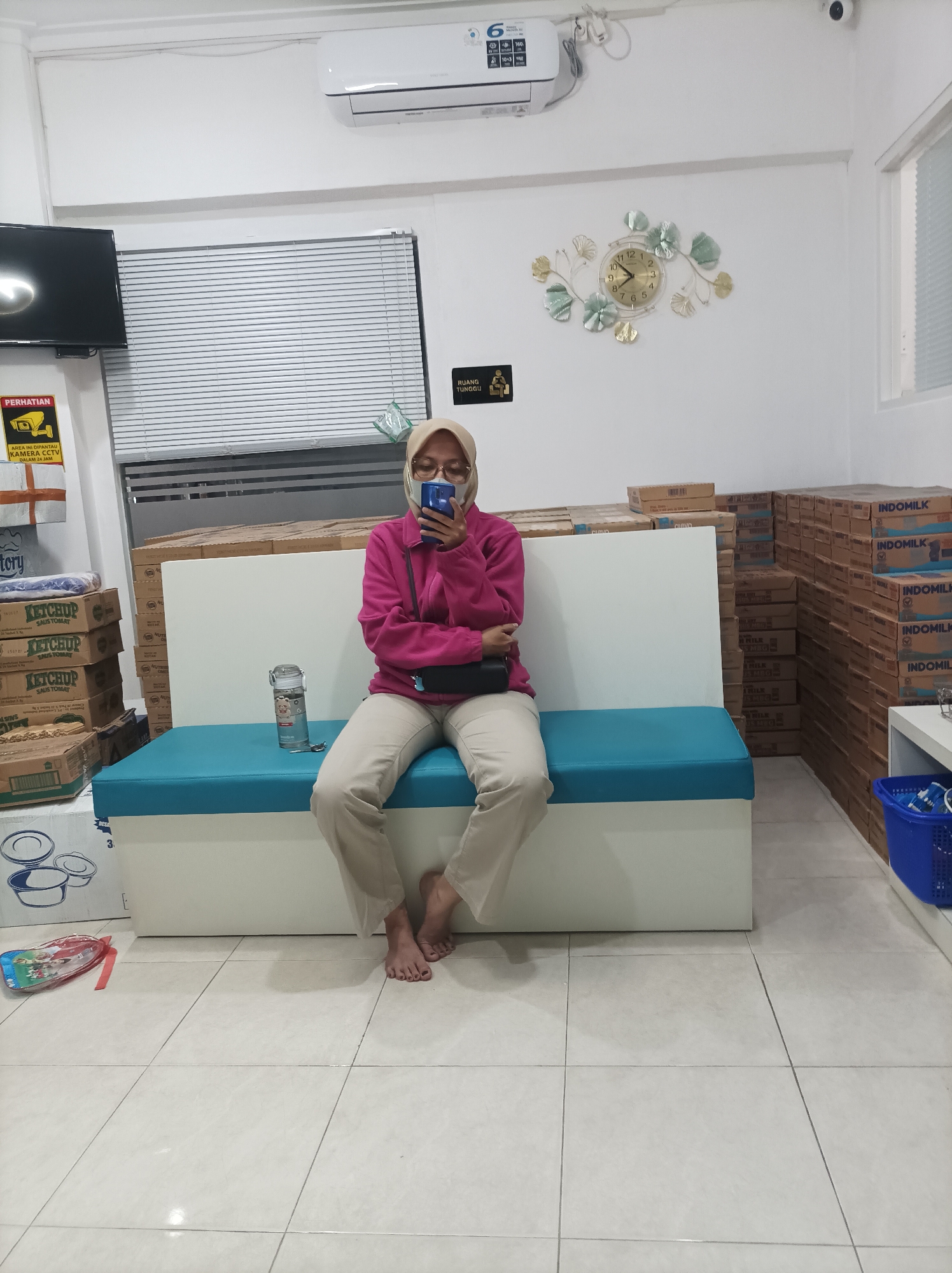 A woman wearing a headscarf and glasses, a pink jacket, and tan pants is sitting on a turquoise bench in what appears to be a waiting area. She is holding a smartphone and looking at it. There are stacks of boxes to her left and right, with one box on the left labeled "Ketchup". A clock is on the wall behind her. To the left of the bench, on the floor, is a colorful children's backpack. The floor is tiled. The scene is indoors, with what looks like an air conditioning unit mounted on the wall above the woman. There is also a sign on the wall that reads "RUANG TUNGGU" which translates to "WAITING ROOM". The location is identified as Mlilir, Indonesia.