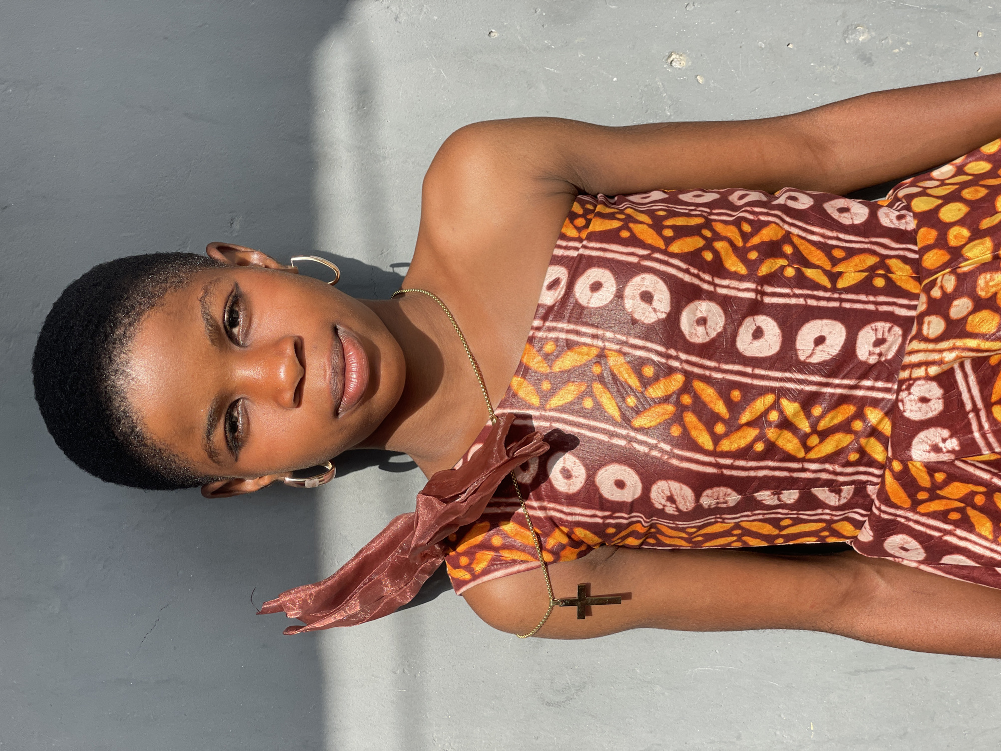 A young woman with short, dark, natural hair is depicted lying down horizontally, looking directly at the viewer with a calm expression and a slight, soft smile. She wears gold hoop earrings and a thin gold chain necklace with a prominent cross pendant that rests on her right arm. Her attire is a strapless garment, likely a top or dress, made from a vibrant patterned fabric. The fabric features a rich maroon or dark brown base adorned with distinct yellow, orange, and white patterns, reminiscent of traditional African prints. A sheer, rust-colored fabric detail is draped around her left shoulder.
The woman is positioned on a plain, textured, light gray surface, which appears to be concrete or a similar outdoor ground material. The scene is bathed in strong, direct sunlight, creating sharp, diagonal shadows across the background, indicating bright, sunny weather during daytime, likely late morning or early afternoon. This lighting highlights the texture of the surface and the woman's skin. The minimalist background focuses attention on the subject. The setting is consistent with an outdoor location in Badagry, Nigeria, known for its bright sun. There is no visible text in the image.