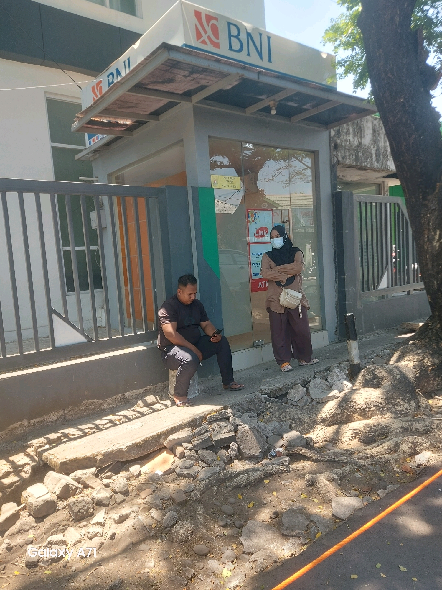 At a BNI bank branch in Makassar, Indonesia, a man sits on a concrete block, looking at his phone. A woman in a hijab and face mask stands nearby, arms crossed, looking towards the bank's entrance. The area appears to be under construction or repair, with debris and broken concrete scattered on the ground. A large tree trunk is visible on the right side of the image.
