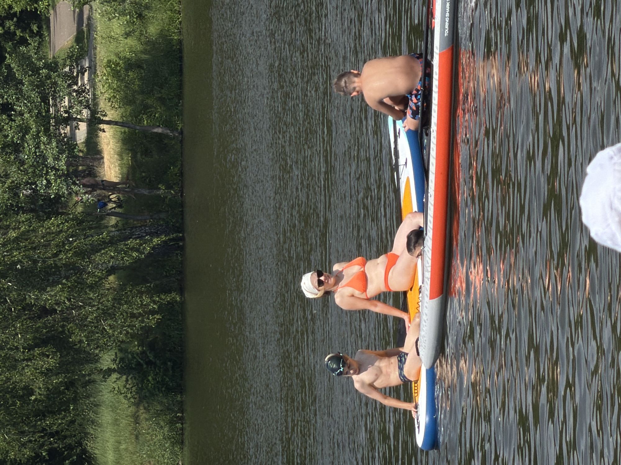 The image depicts three individuals paddleboarding on a body of water, possibly a lake or river, with a lush green shoreline visible to the left. The sun appears to be shining, casting reflections on the water, suggesting it is daytime. Two individuals are positioned on one paddleboard, with one lying down and the other seated. A third person is on a separate paddleboard further down the water. All three individuals are dressed in swimwear. The person lying down on the first paddleboard is wearing an orange bikini and sunglasses, and has a headscarf on. The person seated on the same paddleboard is a young male wearing swim trunks and a swim cap. The third person, also a young male, is wearing swim trunks and a swim cap. The paddleboards are colorful, with red, white, yellow, and blue elements. The image is oriented vertically. Text visible on one of the paddleboards reads "TOURING SHARK". The scene appears to be a recreational activity on a warm, sunny day, with the participants enjoying themselves on the water. The lush vegetation on the shore suggests a natural, outdoor setting.