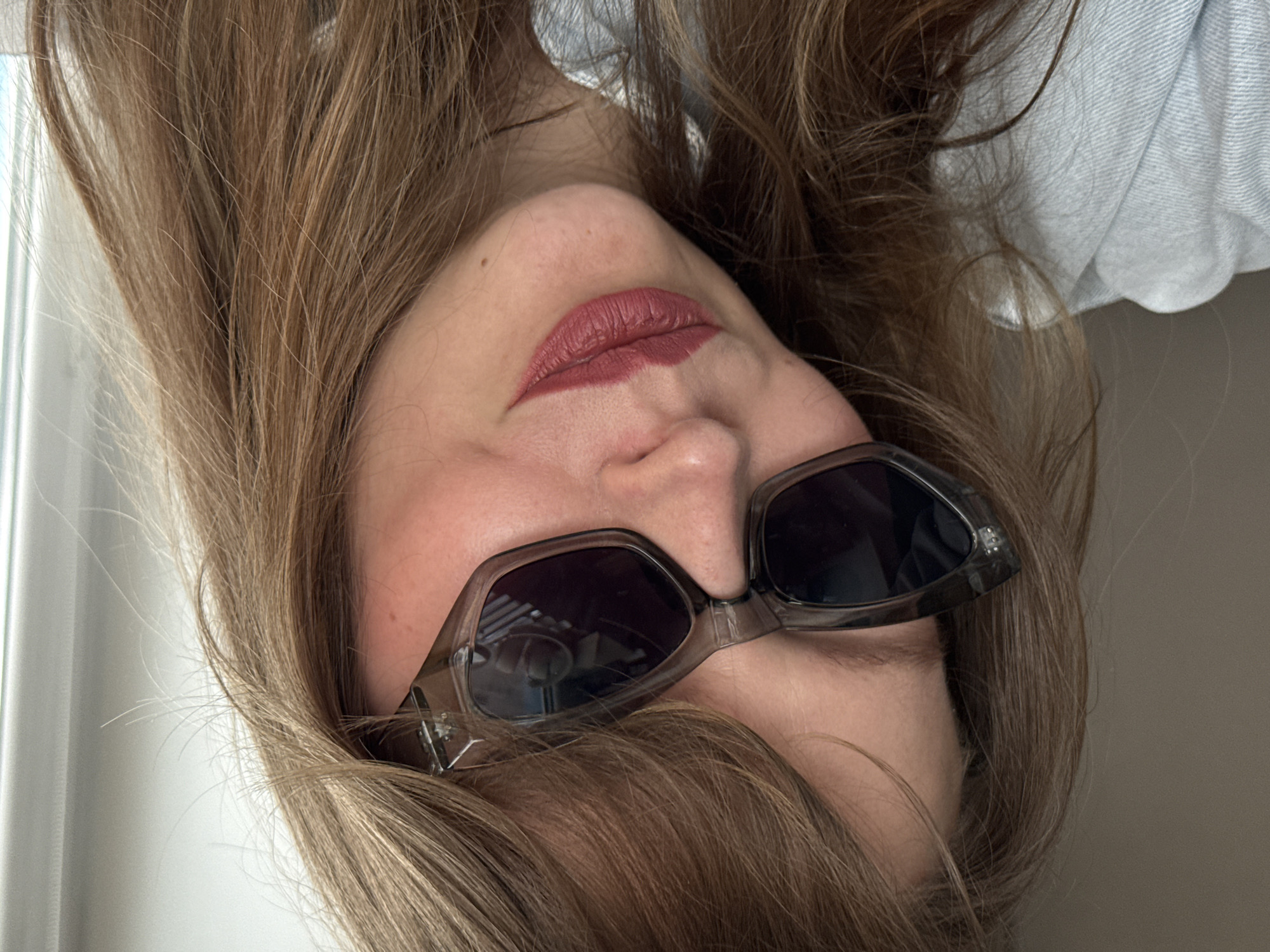 A woman with light brown hair is wearing sunglasses and lipstick. She is wearing a light blue shirt. The image is rotated upside down. The location is not evident from the image and cannot be determined without additional information.