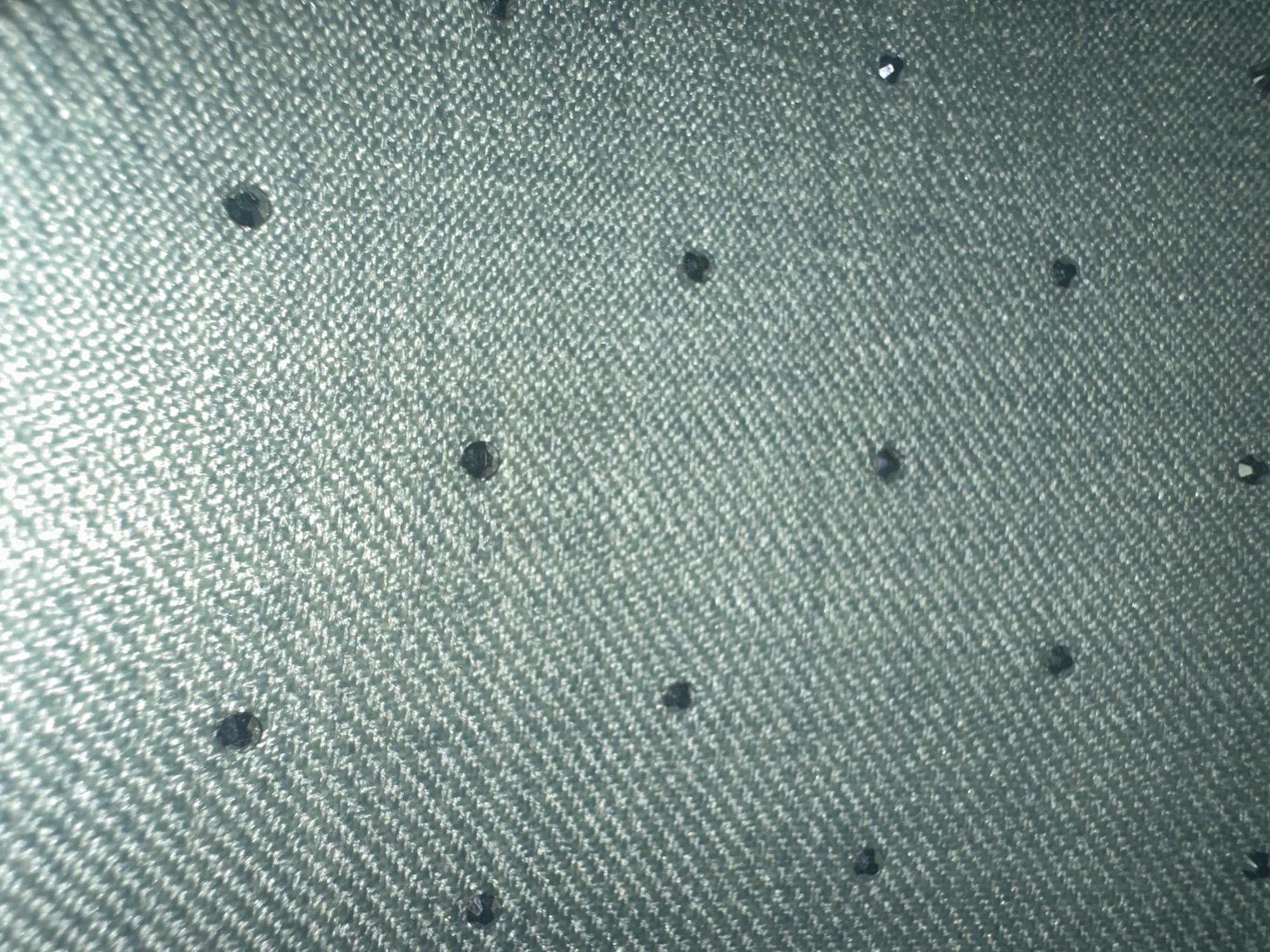 This media file is a close-up photograph of a textured, greenish-blue fabric, possibly denim, adorned with small, dark, faceted embellishments that appear to be rhinestones or beads. The fabric has a distinct woven pattern, with diagonal lines of raised texture. The embellishments are scattered unevenly across the surface, with some appearing more prominent and reflective than others, suggesting they catch the light. The lighting in the image is somewhat harsh, creating bright highlights on the fabric and the embellishments, and casting subtle shadows. The overall impression is of a decorative textile, likely a garment or accessory, viewed from a macro perspective. There is no discernible text, people, or specific environmental cues to suggest a particular time of day, weather, or outdoor location. The context of Letterkenny, Ireland, is provided externally, but the image itself does not contain visual information to confirm this setting.