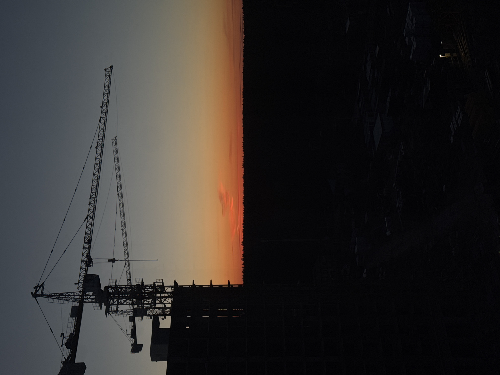 The image depicts a silhouette of construction cranes and a building under construction against a dusky sky, likely at dusk or dawn. The sky fades from a light grey to orange near the horizon, suggesting the presence of a sunset or sunrise. To the right, a dense dark area suggests a forest or a heavily built-up city landscape. The construction site is a complex network of metal beams and cranes, typical of a large building project. The image is rotated, so the original orientation is unclear. The location of the scene is in Izhevsk, Russia.
