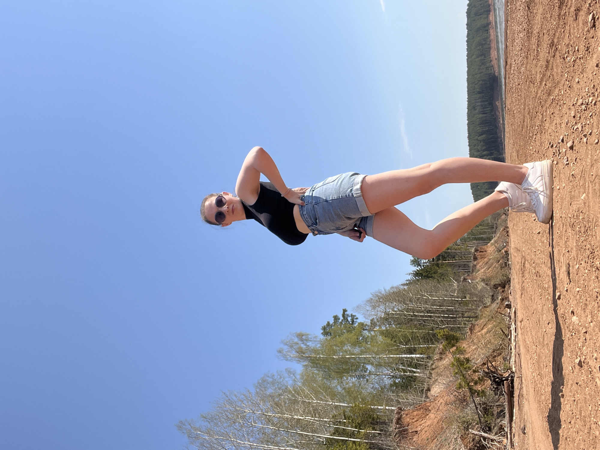 A medium-shot photograph, oriented sideways, shows a young woman standing on a dirt embankment under a bright blue sky.
The woman wears denim shorts, a black crop top, and white sneakers. She is wearing sunglasses and has her hands on her hips.
The background consists of trees below the embankment and a dirt path with a line of coniferous trees in the distance above the embankment. The location is Камское, Russia.
