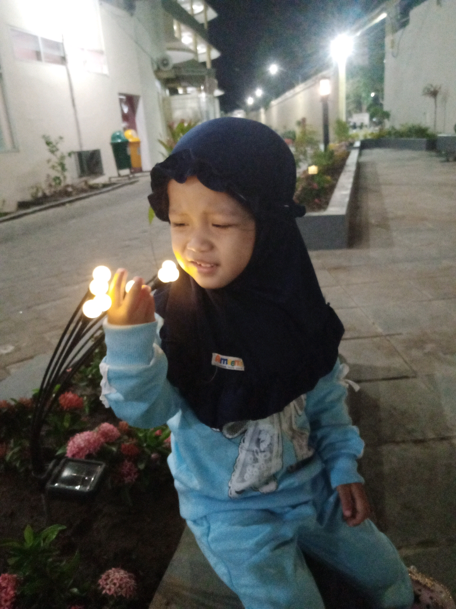 In the city of Kediri, Indonesia, a young child wearing a dark blue hijab and a light blue outfit with a graphic print on the chest, is captured in a nighttime scene. The child is reaching out a hand towards a string of small, glowing lights. The background shows a paved walkway, a garden area with pink flowers and a solar-powered light, a white building, and streetlights illuminating the scene. The overall impression is one of a child's wonder and curiosity in a softly lit outdoor environment.